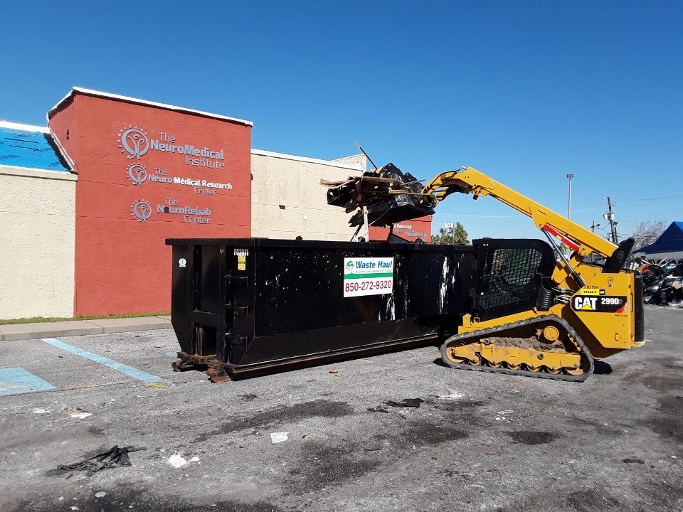 Yellow excavator loading debris into a black dumpster beside a red building under a clear blue sky
