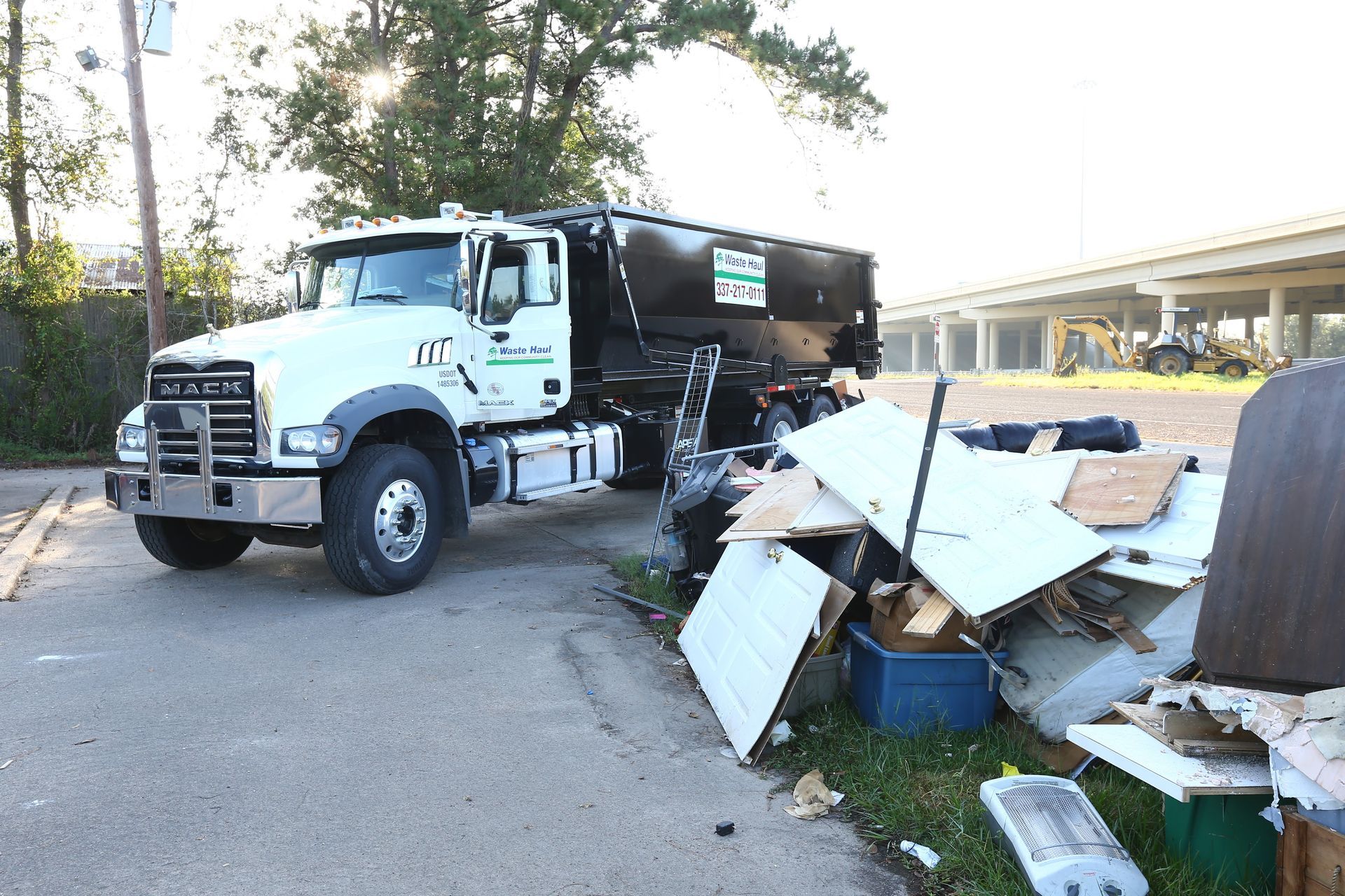Dump truck beside a roadside pile of debris and discarded furniture under an overpass