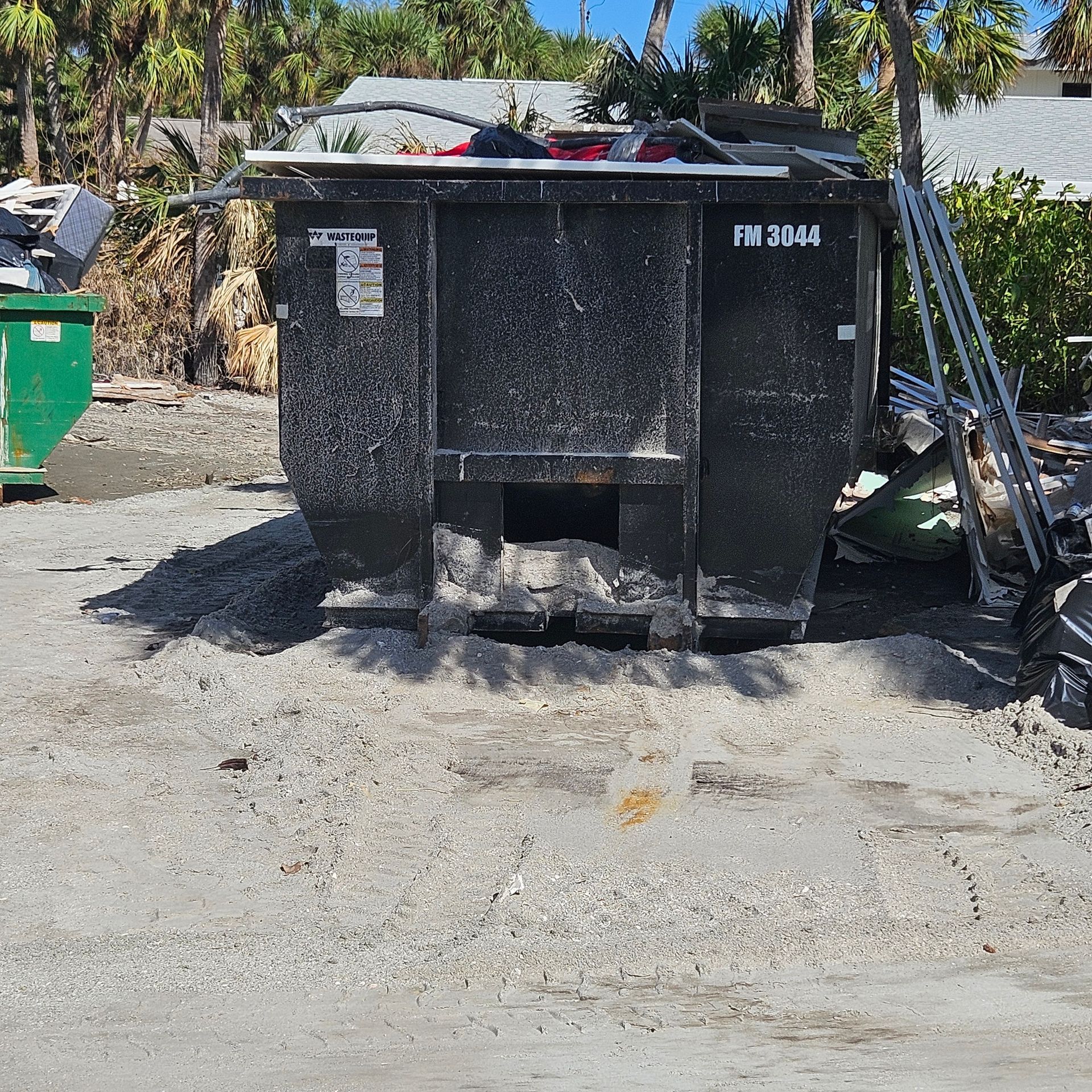 Large black dumpster on a sunlit dirt lot, with trash bins and debris nearby.