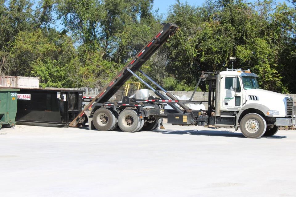 White tow truck with raised boom beside a dumpster in a sunny outdoor lot