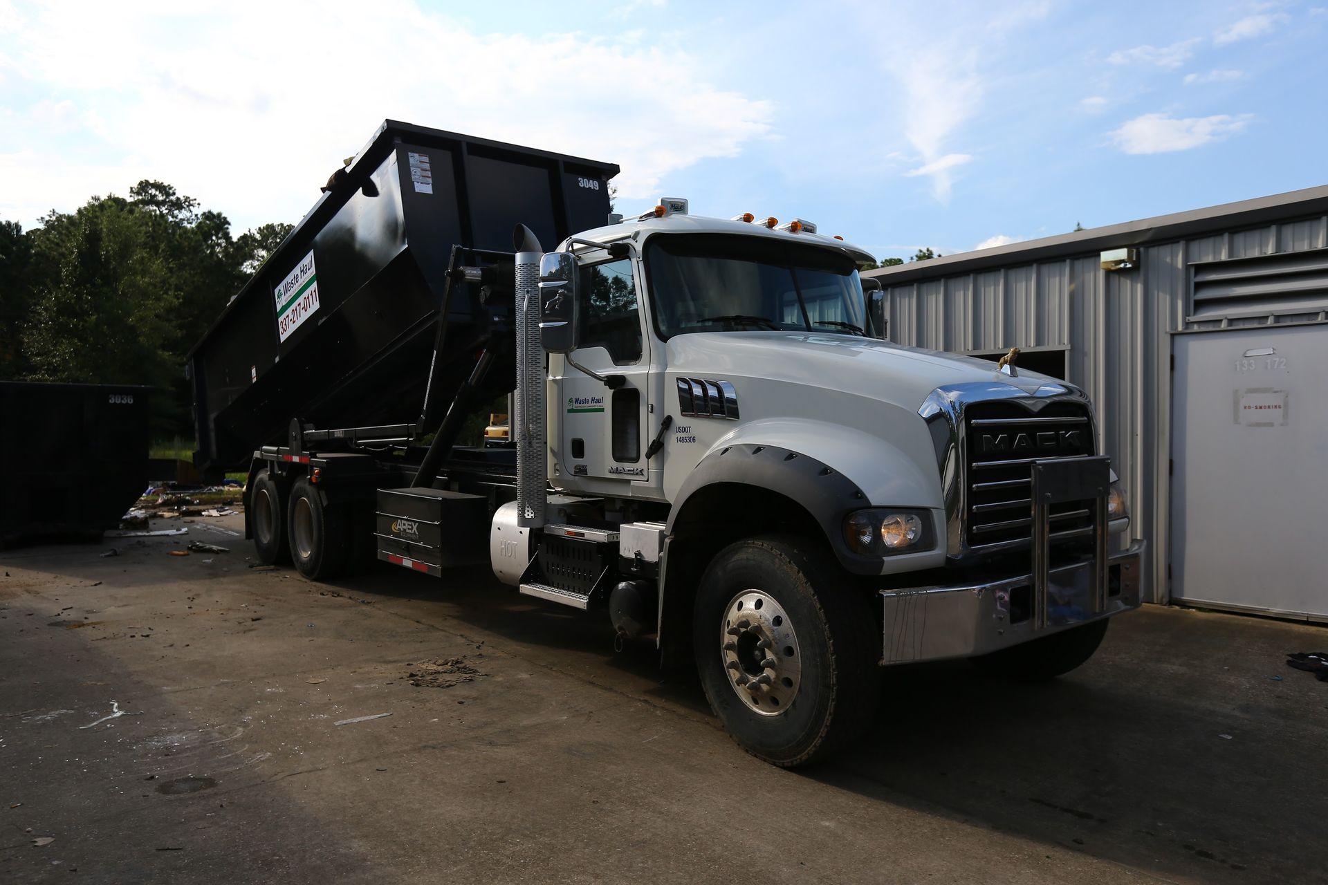 White dump truck parked beside a building, carrying a large black container.