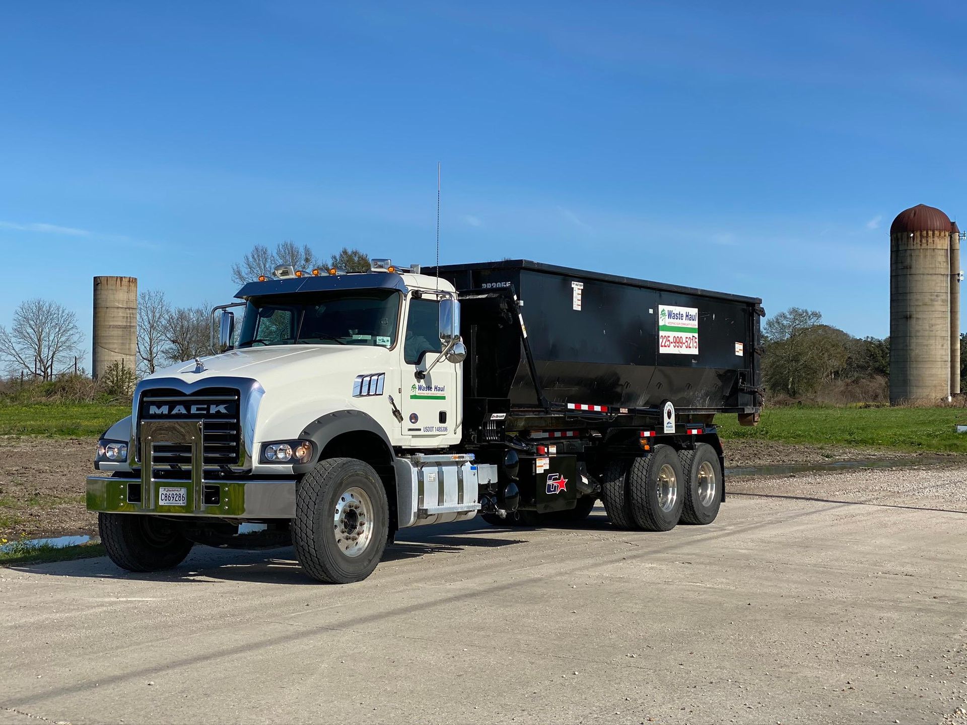 White and black dump truck parked on a paved lot with grassy field and silos under a blue sky