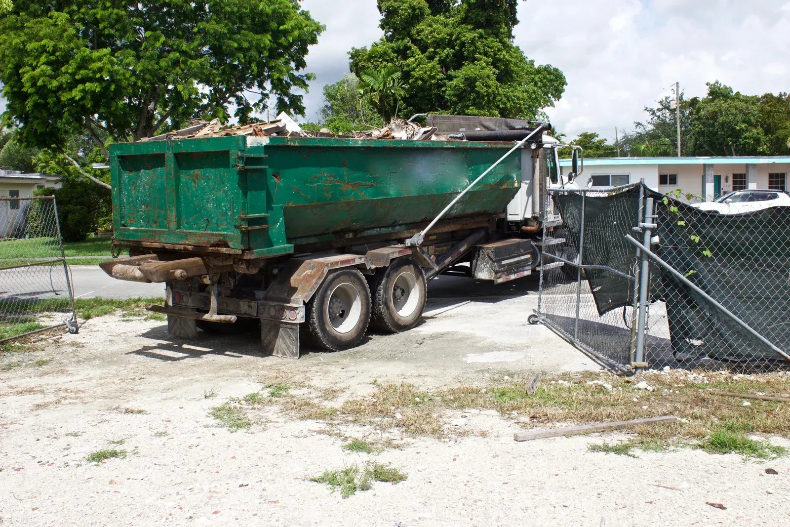 Green dump trailer parked beside a chain-link fence in a gravel yard.