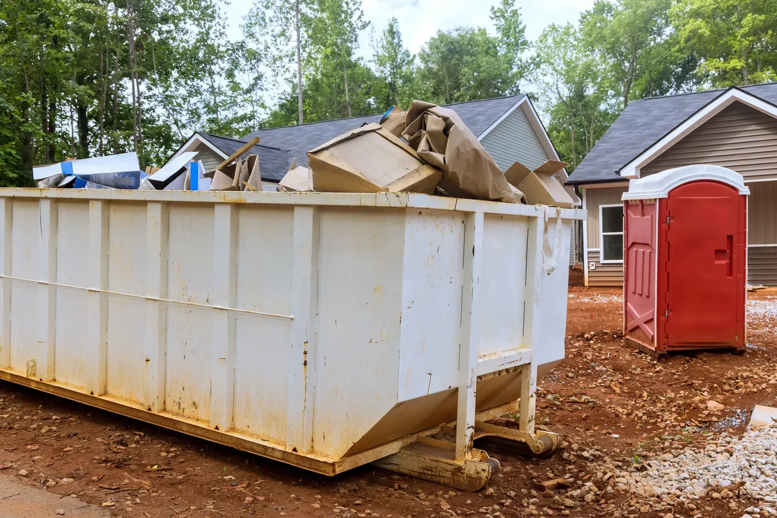Large white dumpster filled with debris beside a red portable toilet at a construction site