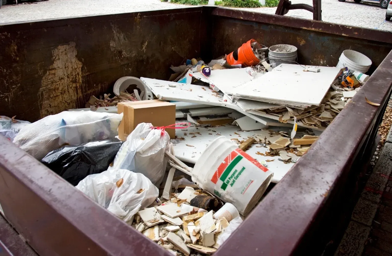 Dumpster filled with construction debris, broken white panels, and plastic bags