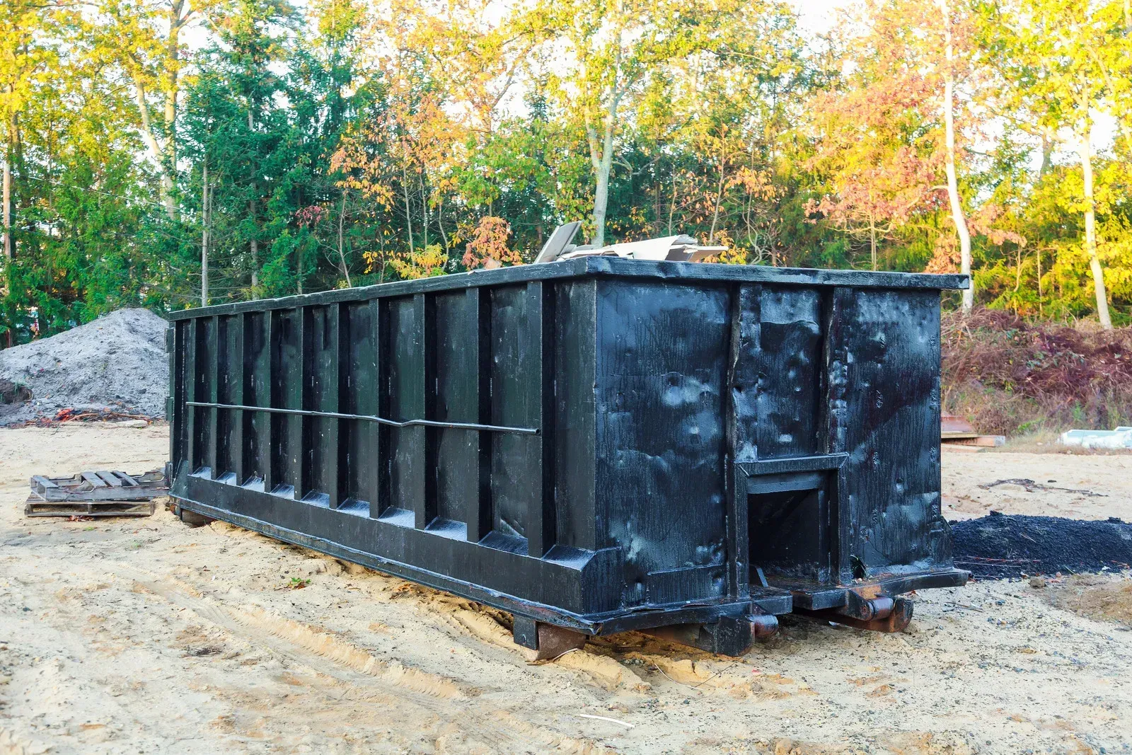 Black construction dumpster on a dirt lot with trees in the background