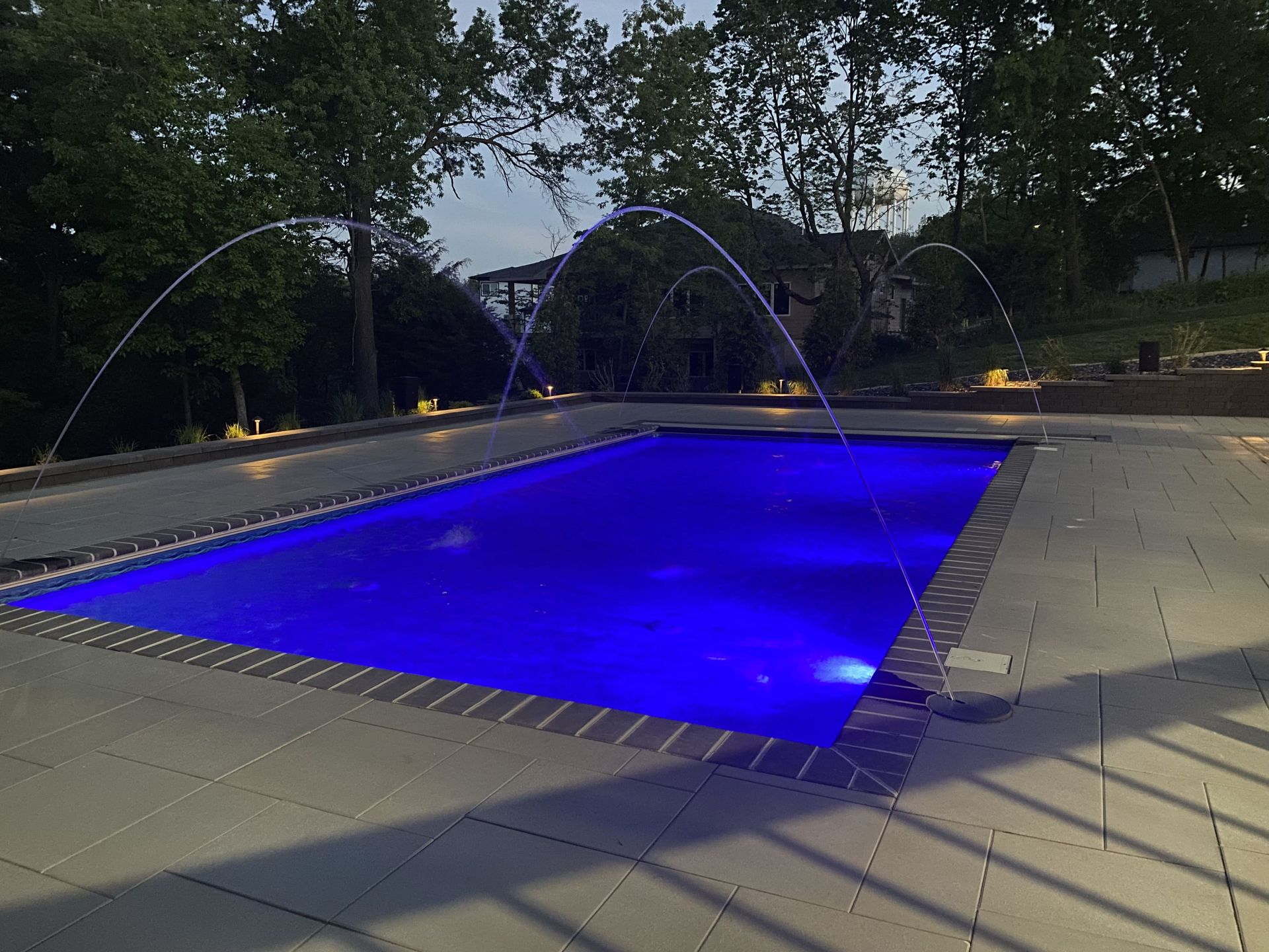 Illuminated blue swimming pool at night with water jets arching over it and trees around the deck