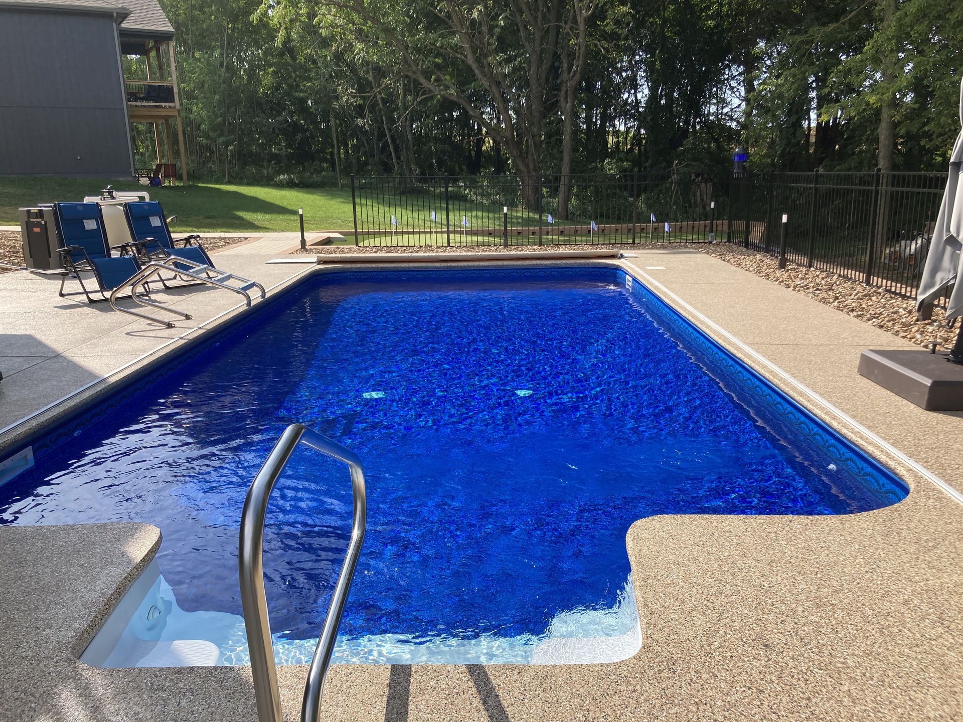 Backyard swimming pool with blue water, patio chairs, and a metal handrail on a sunny day
