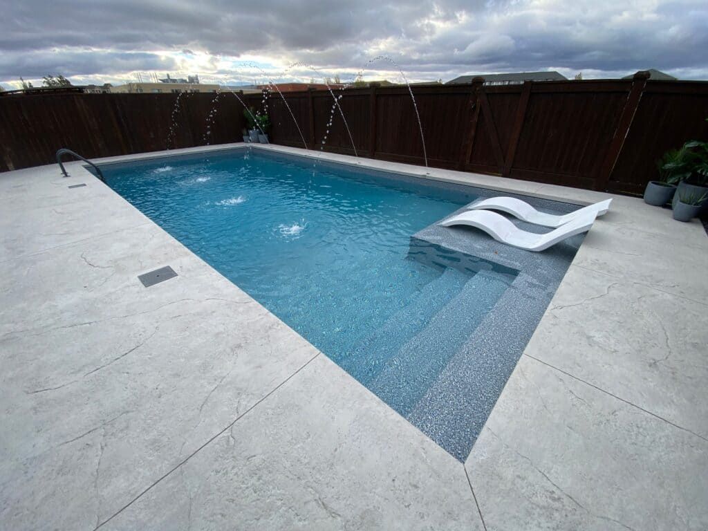 Backyard pool with gray deck, dark wood fence, and two white lounge chairs by the water