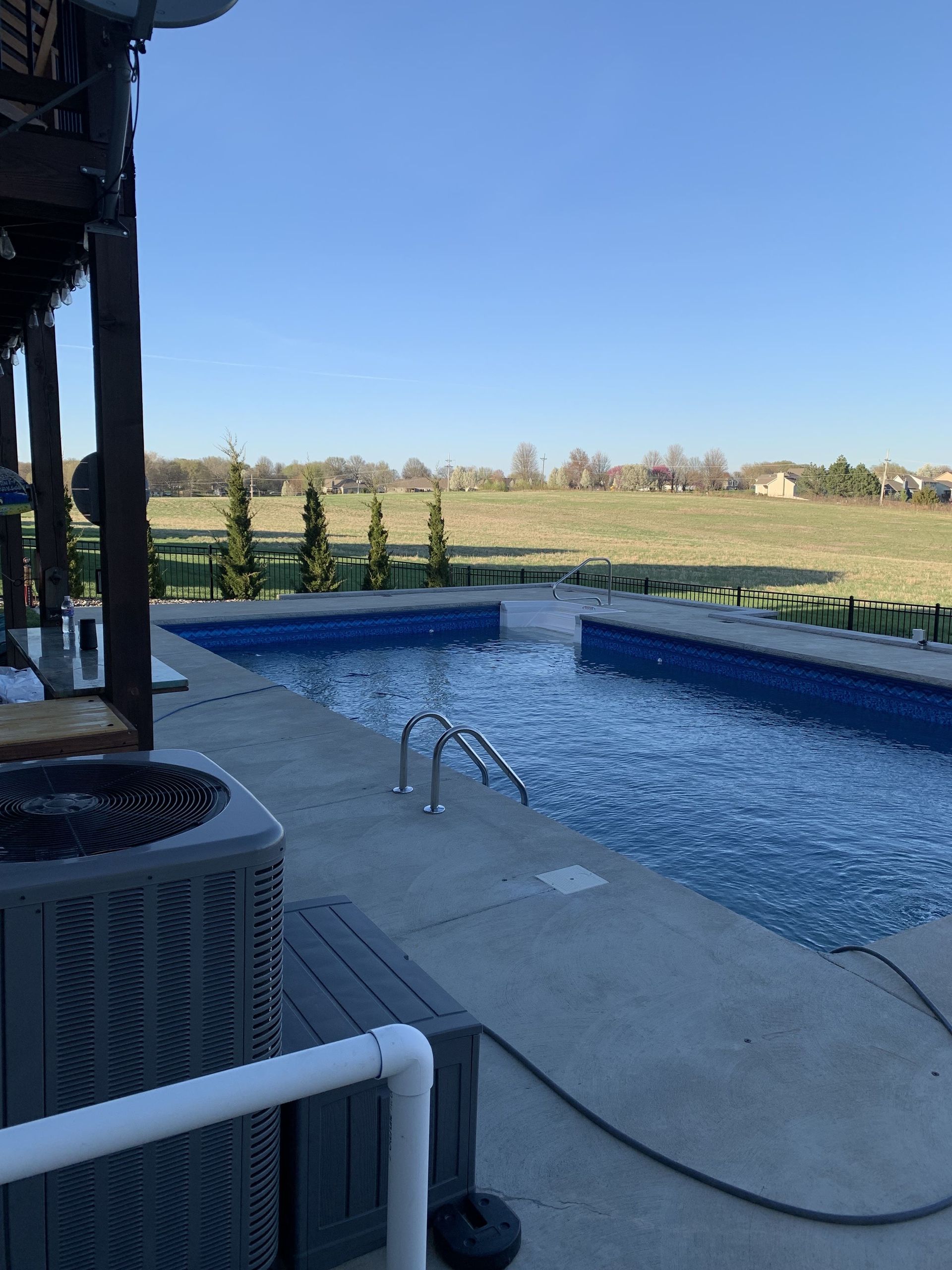 Backyard patio with a swimming pool, hose, and grassy field beyond the fence