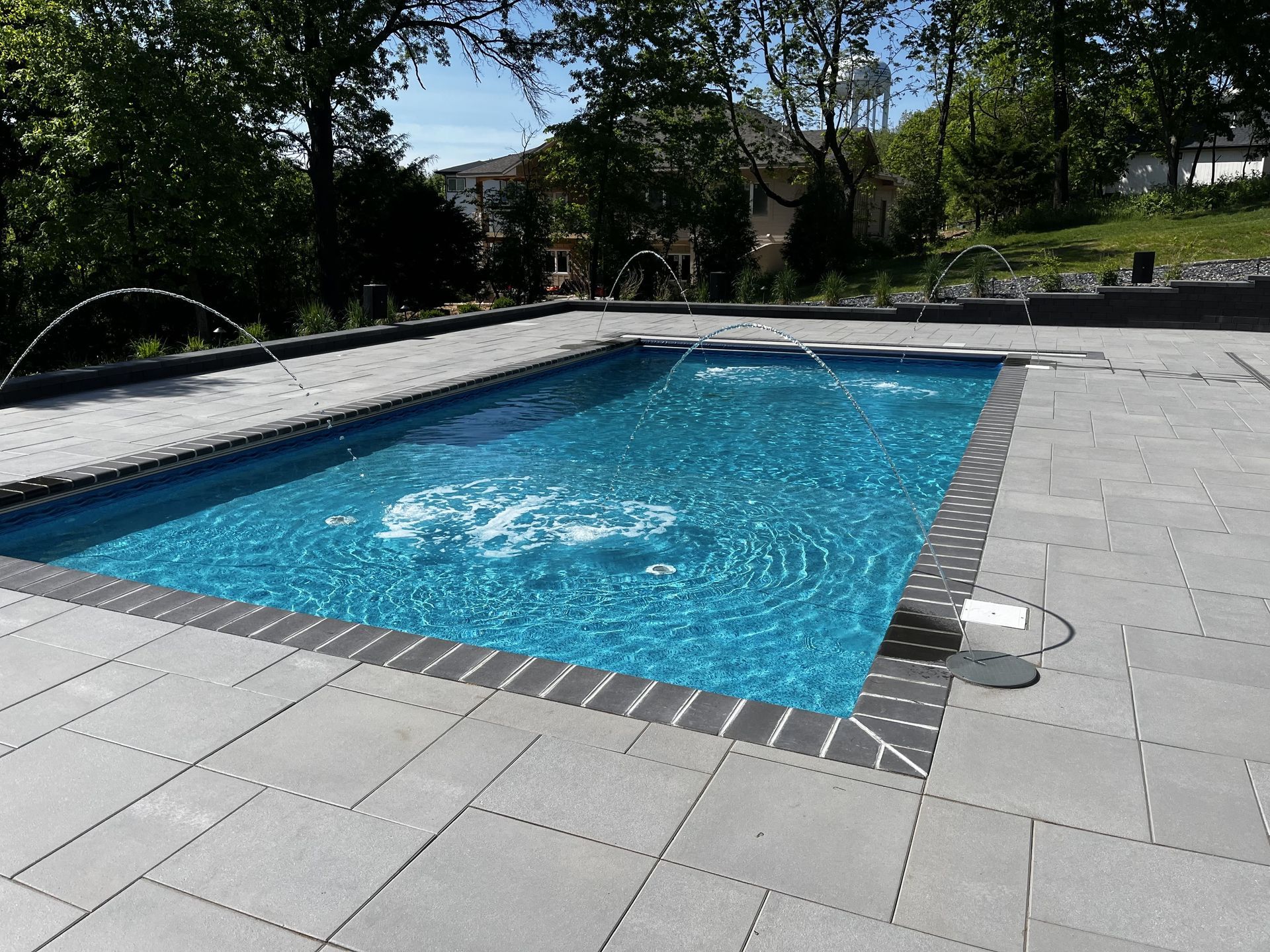 Backyard swimming pool with blue water, gray stone deck, and trees in the background