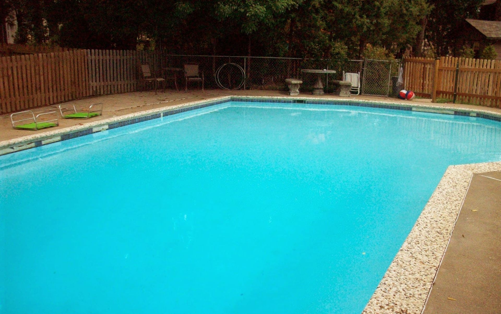 Backyard swimming pool with bright blue water, surrounded by a deck and wooden fence, with trees in the background