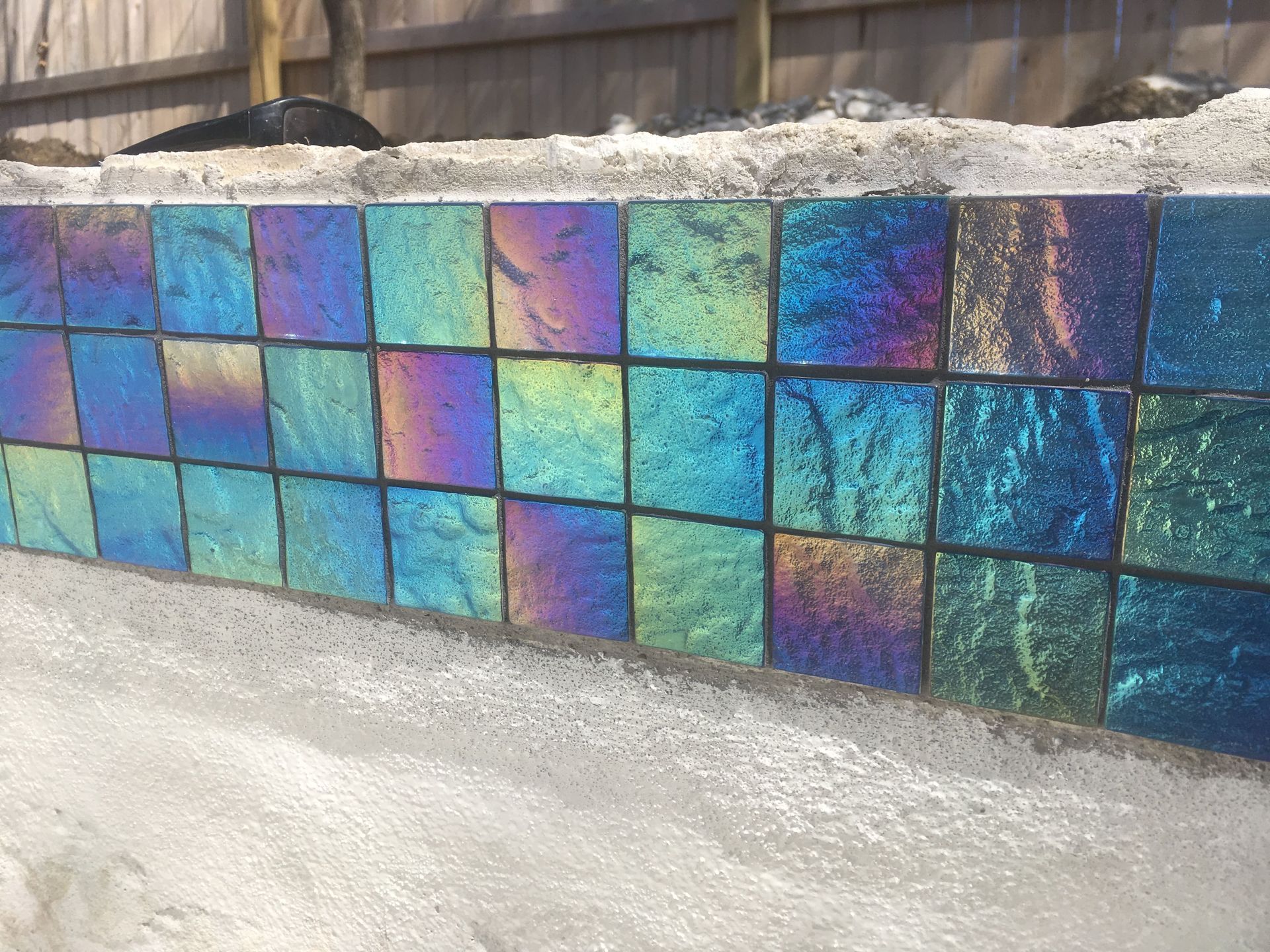 Shimmering blue and purple mosaic tile wall beside a concrete path and wooden fence