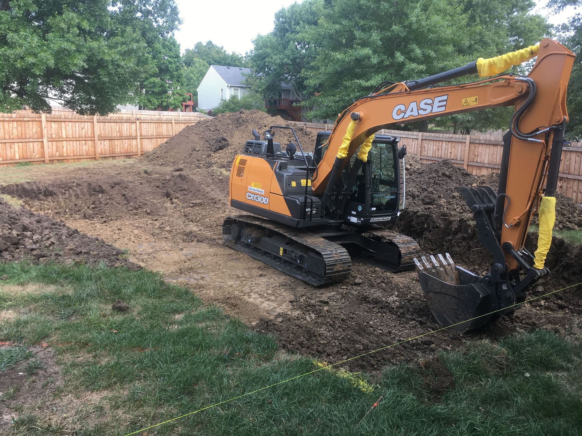 Yellow CASE excavator digging in a dirt backyard beside a wooden fence.