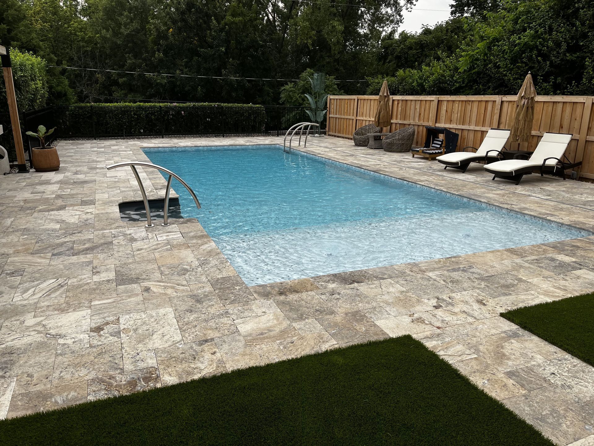 Backyard pool with stone patio, lounge chairs, and wooden fence, surrounded by trees
