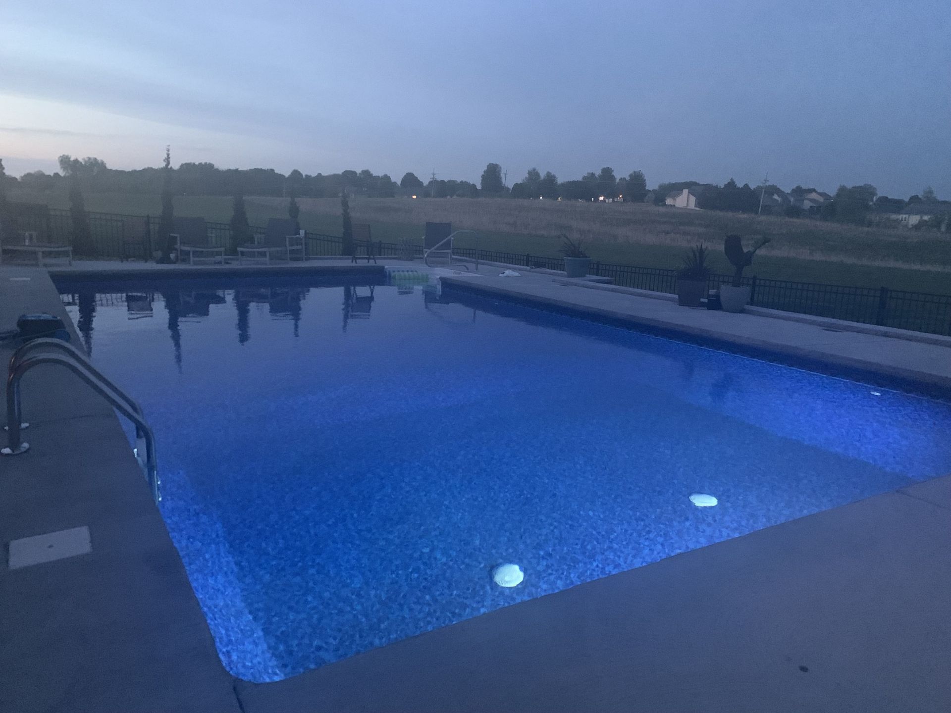 Backyard swimming pool glowing blue at dusk with yard and fence beyond