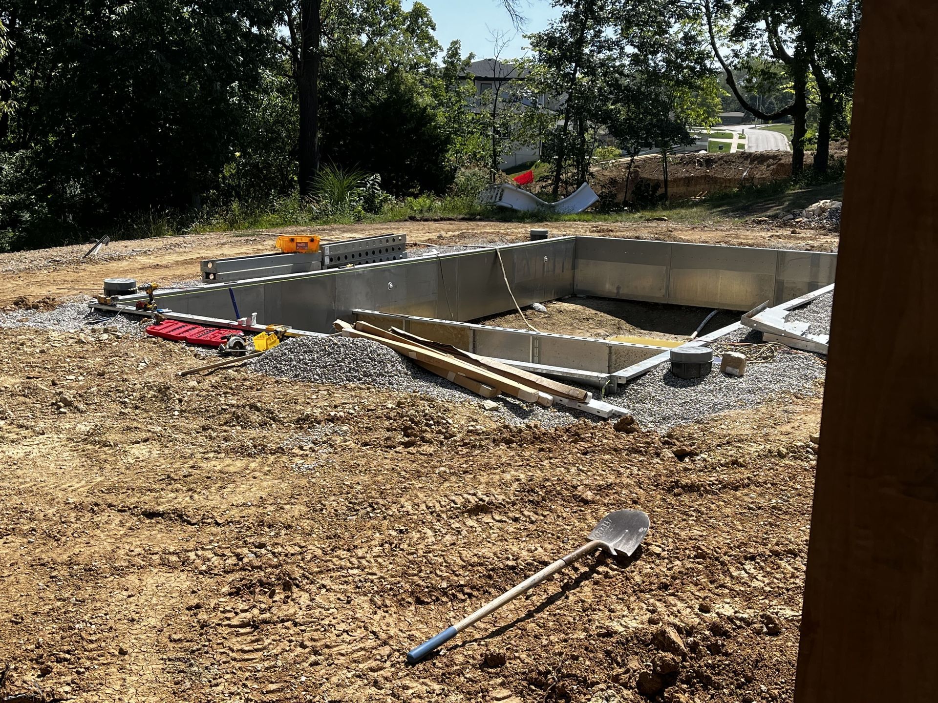 Concrete foundation construction site with wooden forms, gravel, and tools on dirt ground.