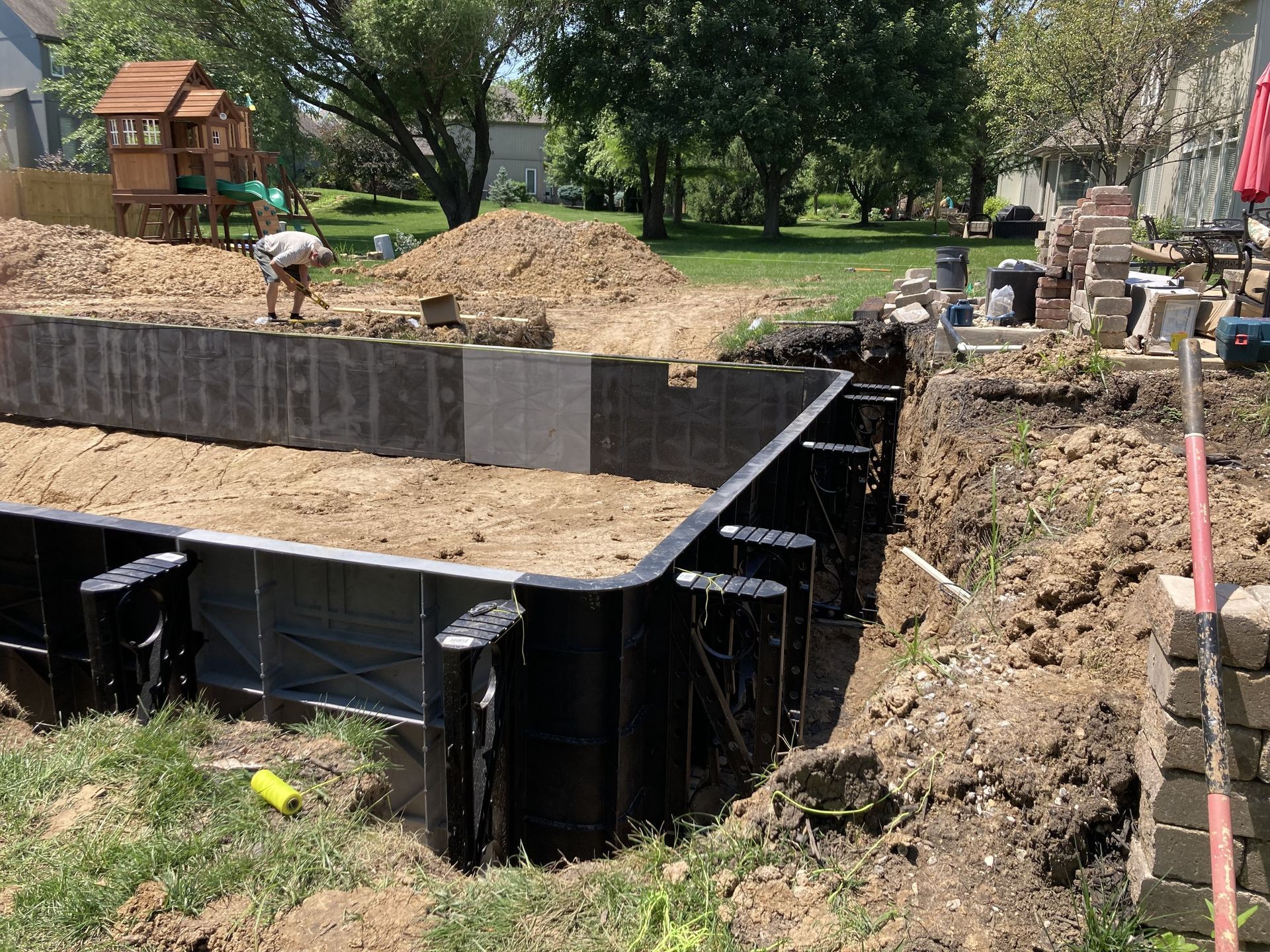 Construction site with excavation pit, black retaining walls, soil piles, and workers in a park-like area