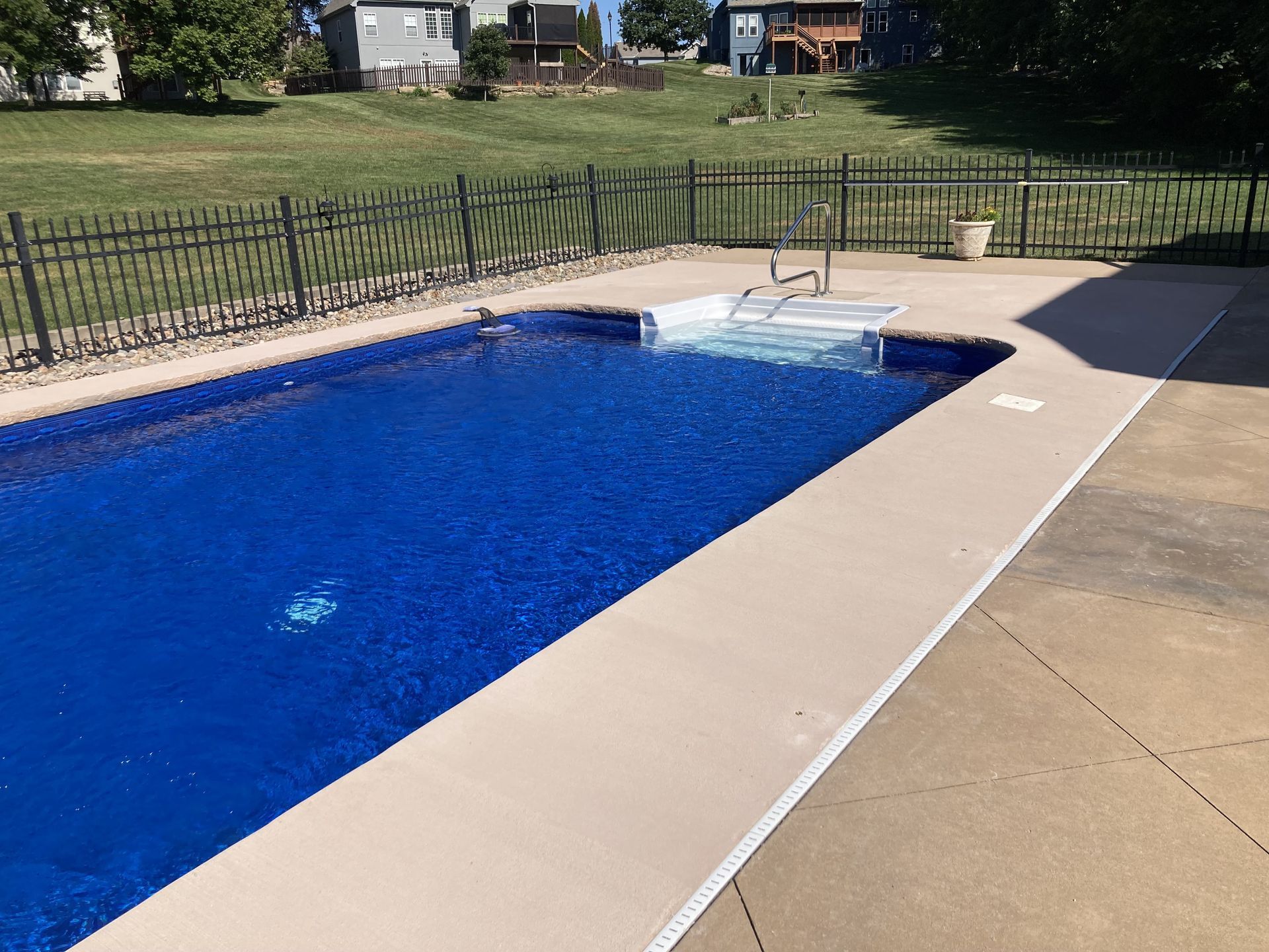Backyard swimming pool with blue water, tan deck, and black fence under sunny skies