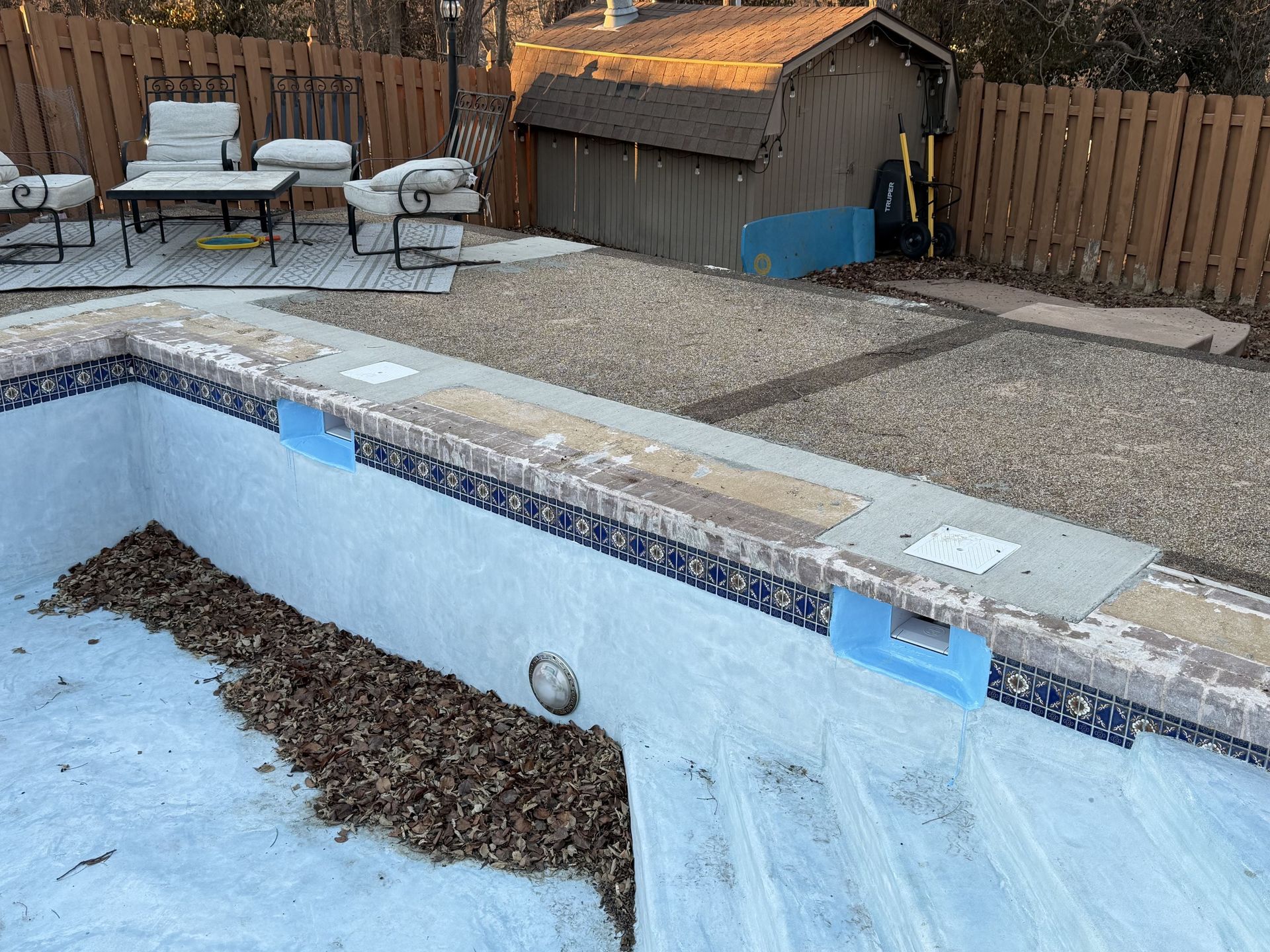 Empty backyard swimming pool with debris, blue tarp, and patio furniture along a wooden fence.