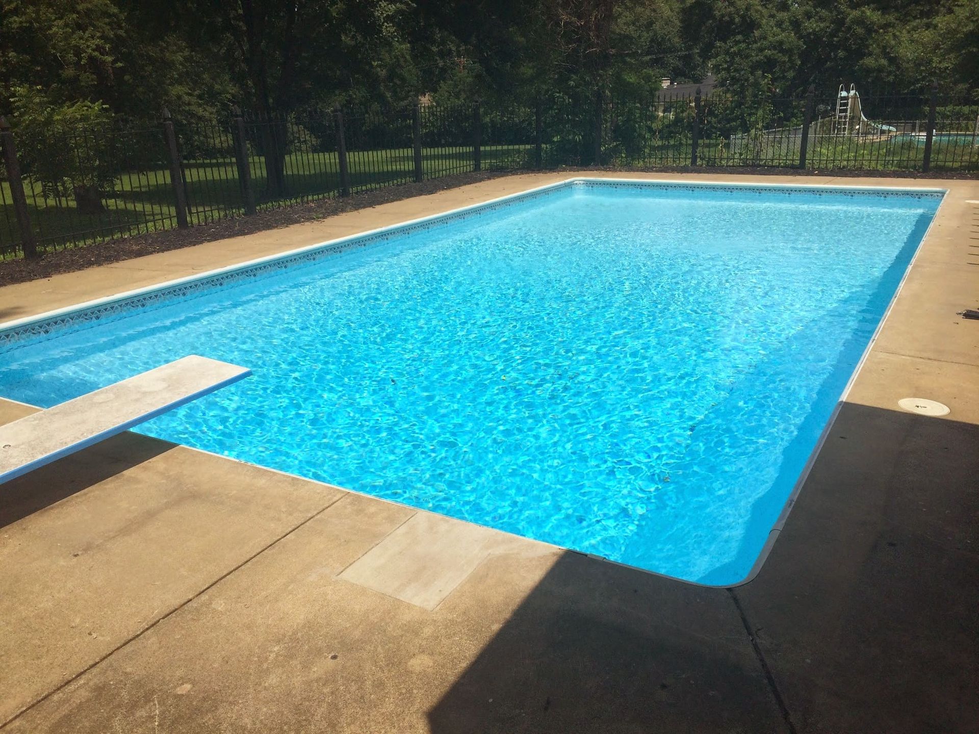 Outdoor swimming pool with clear blue water, tan deck, and surrounding trees and fence