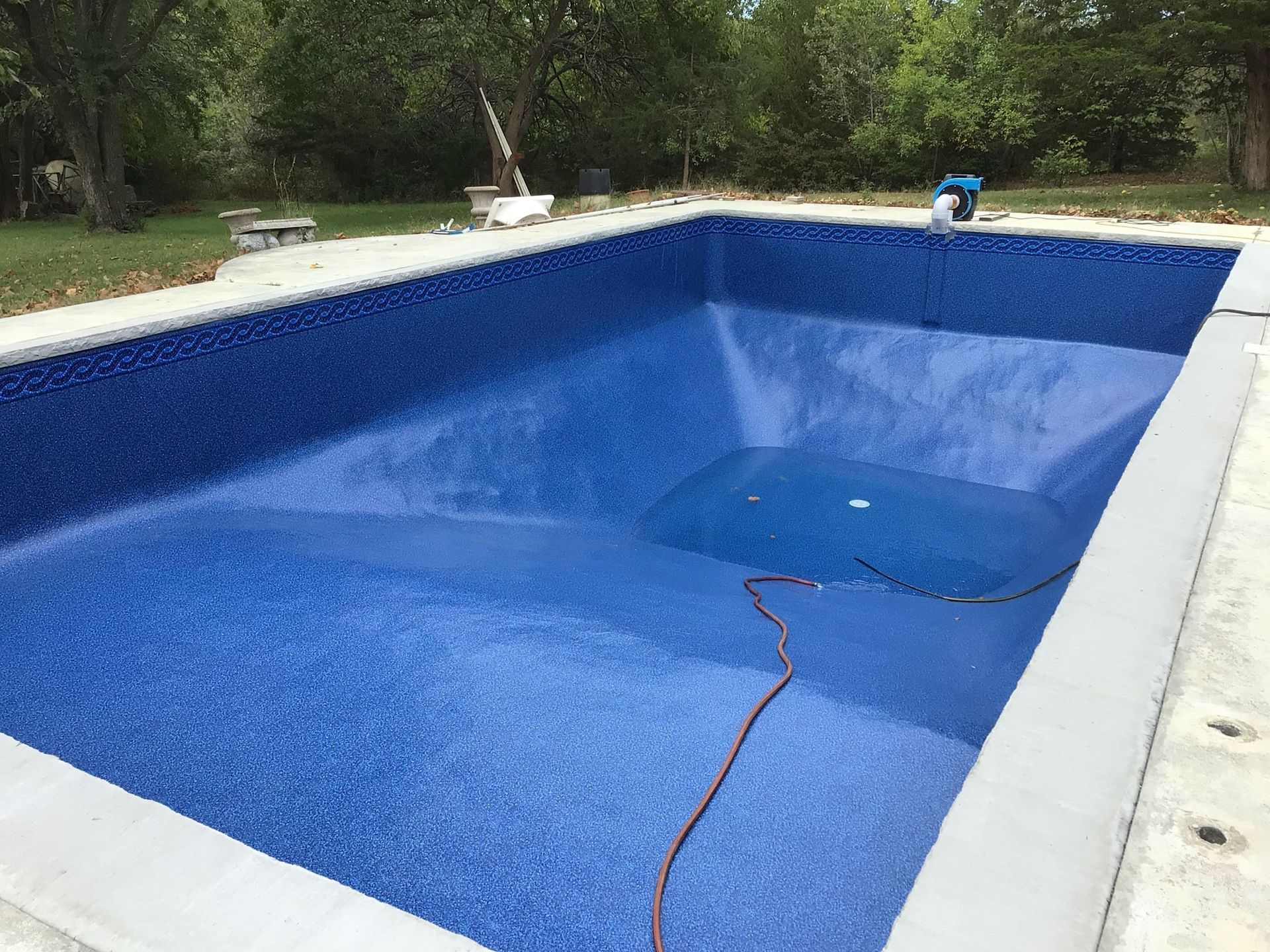 Empty blue in-ground swimming pool with a hose inside and trees around it