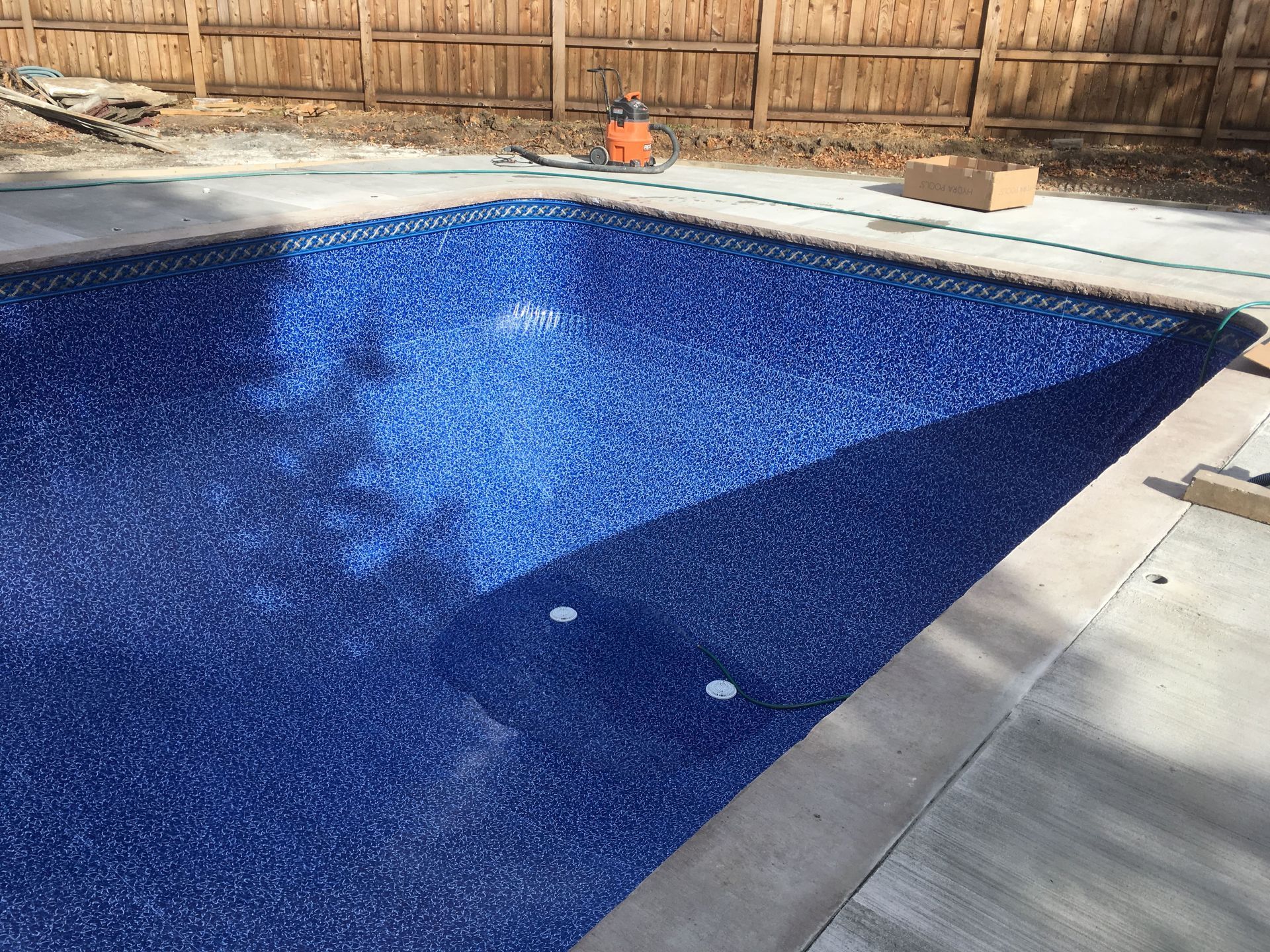 Backyard in-ground pool with blue liner, concrete deck, and wooden fence