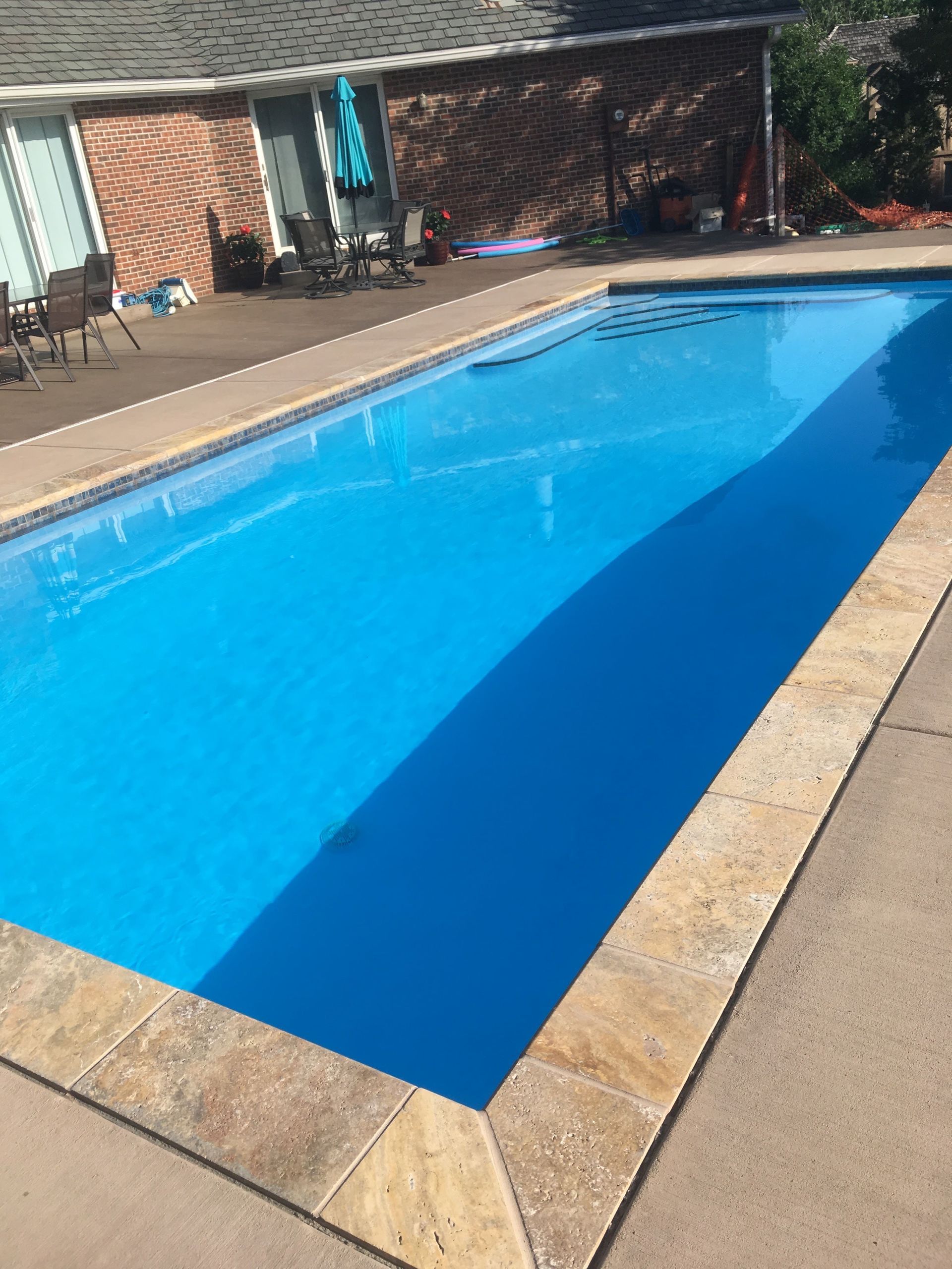 Swimming pool with bright blue water, beige coping, and brick patio beside a shaded lounge area.