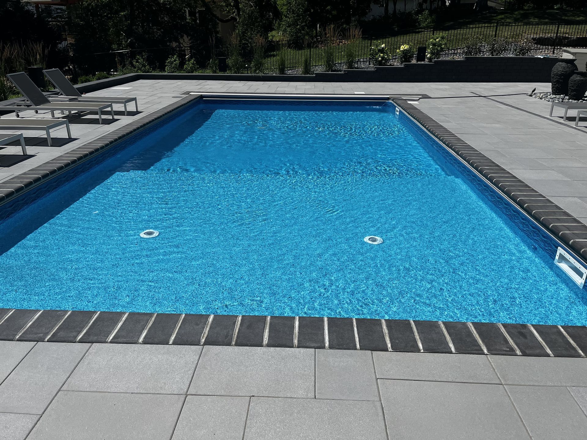 Outdoor swimming pool with bright blue water and gray poolside tiles, viewed from the end.