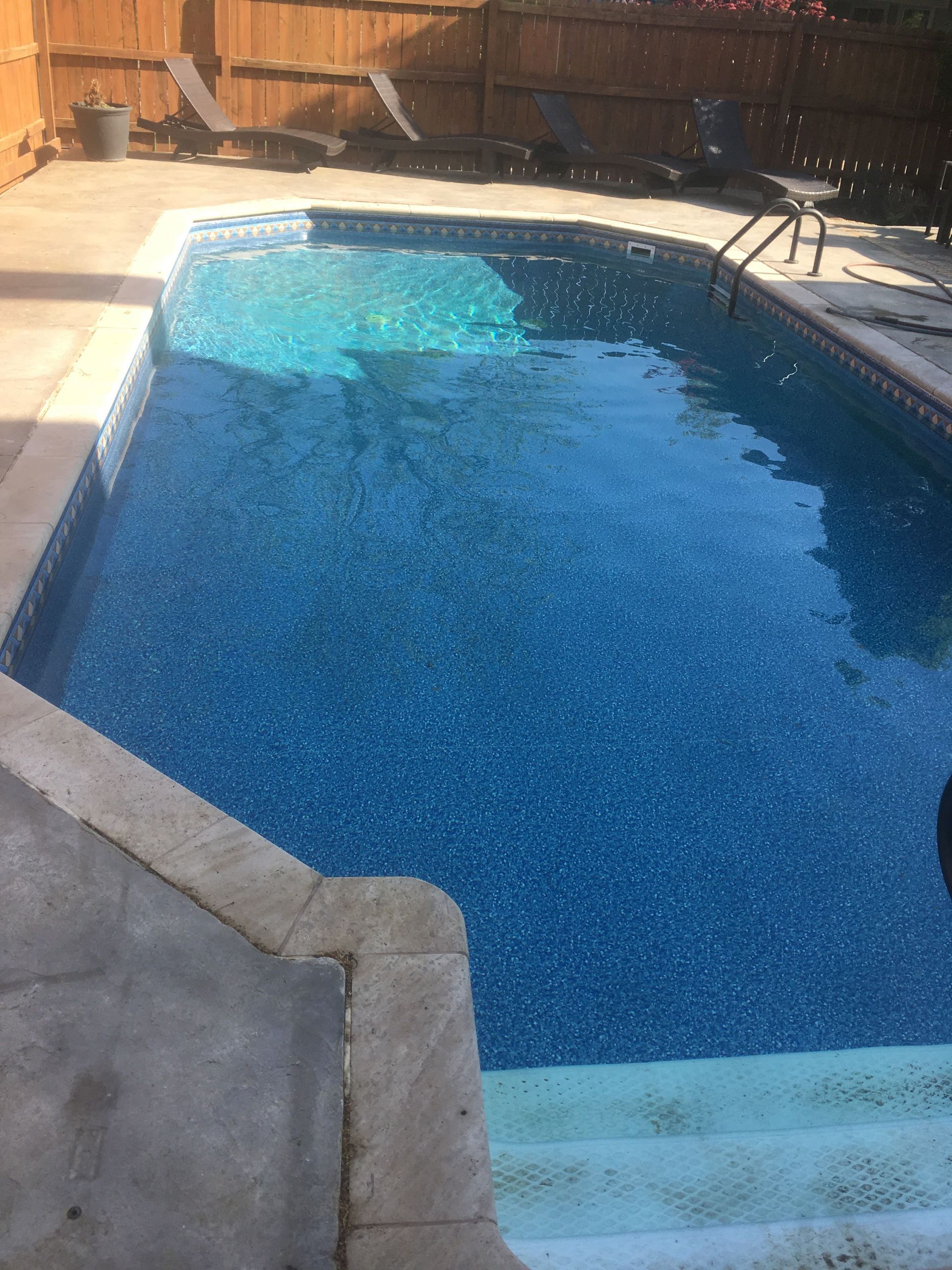 Backyard swimming pool with blue water and concrete deck, bordered by a wooden fence.