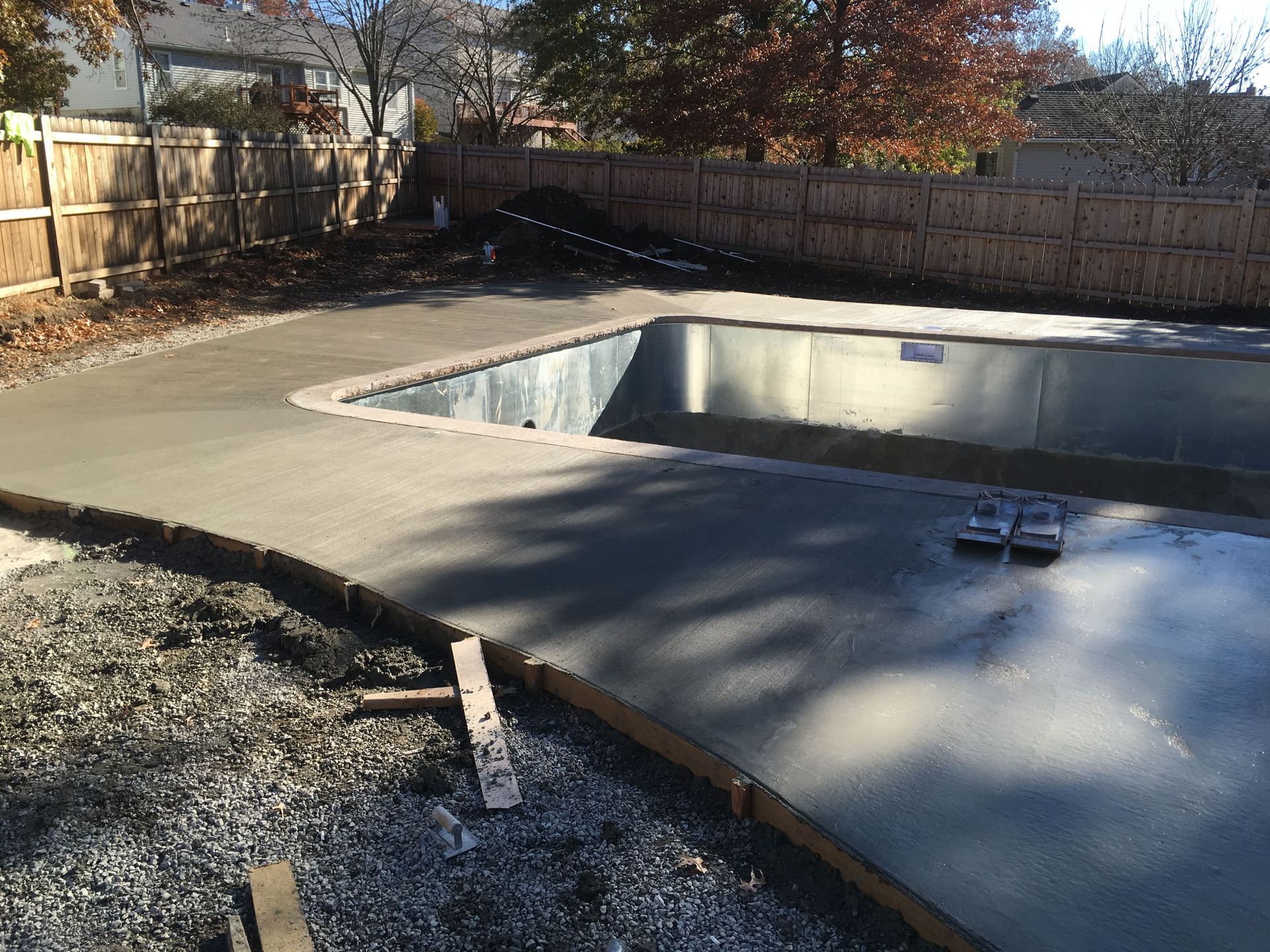 Empty backyard swimming pool surrounded by concrete patio and wooden fence in autumn sunlight