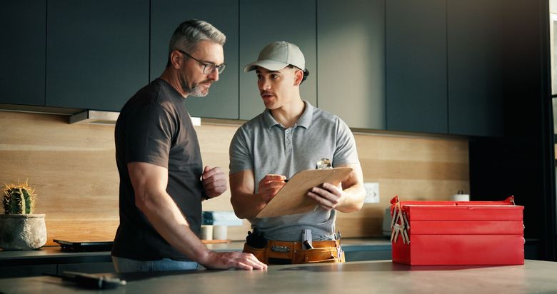 Two men in a kitchen discussing paperwork beside a red tool case on the counter