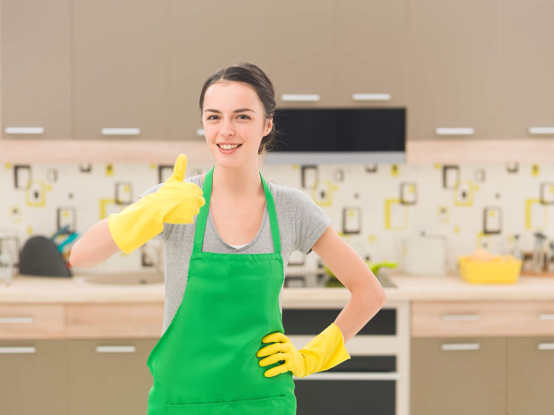 Woman in a green apron and yellow gloves gives a thumbs-up in a kitchen.