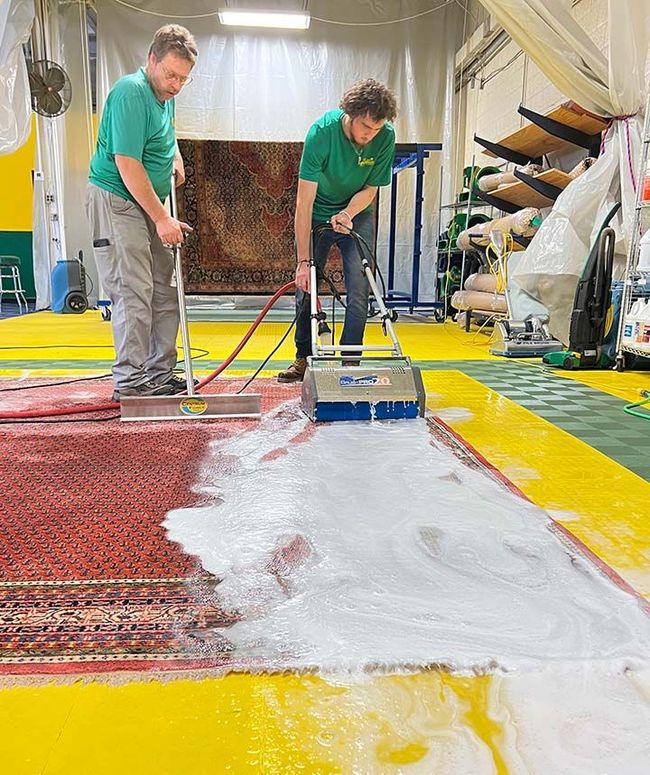 Two people cleaning a rug with machines in a yellow-floored workshop; foam covers part of the rug.