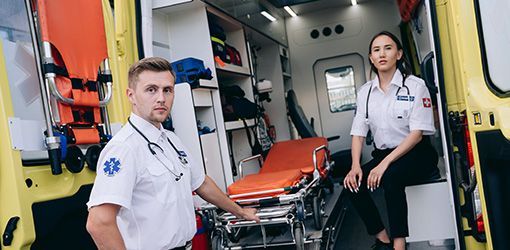 Two paramedics inside an ambulance, one holding the gurney, the other seated, both wearing white uniforms with stethoscopes.