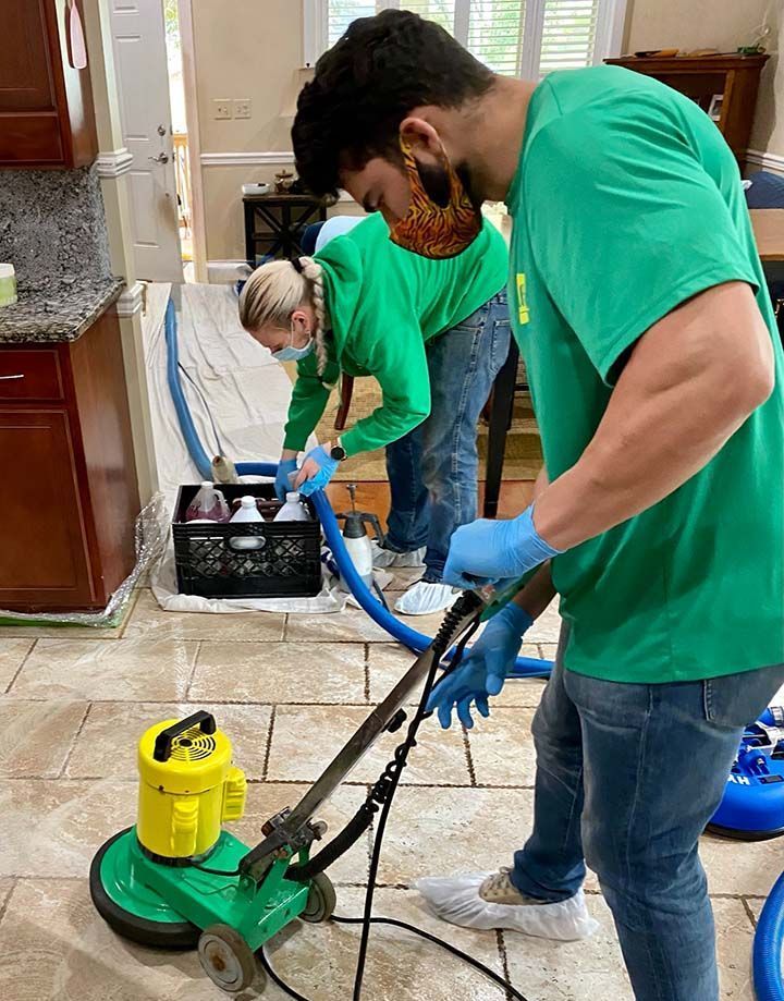 Two people cleaning tile floor with a machine. One operates the machine, the other uses a hose and spray bottle.