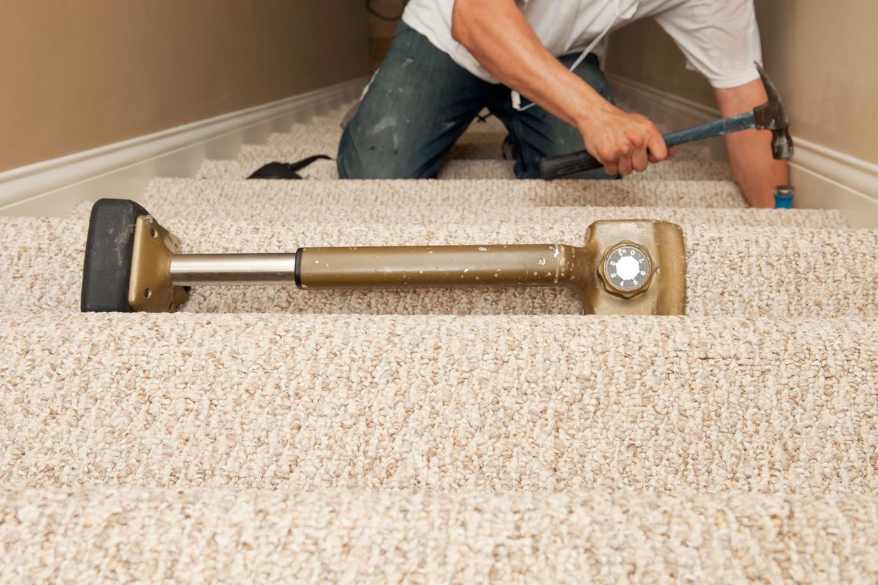 Person installs carpet on stairs with a hammer and stretcher tool. Light beige carpet.