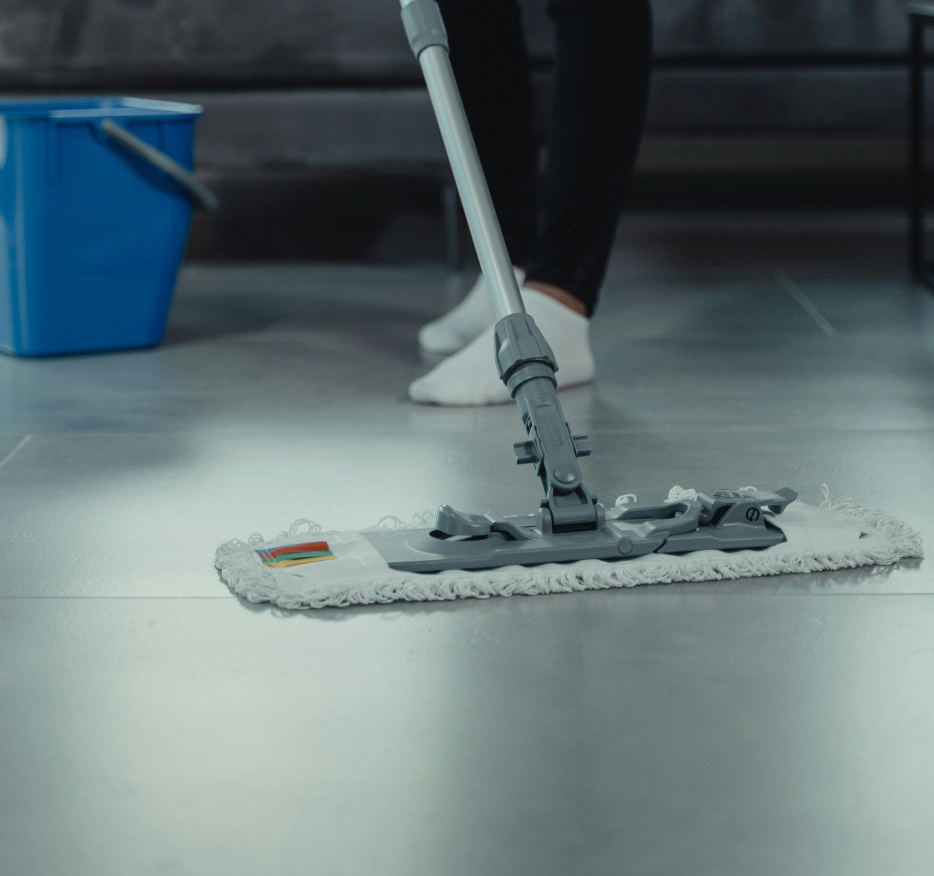 Person mopping a shiny, gray floor with a flat mop; a blue bucket is in the background.