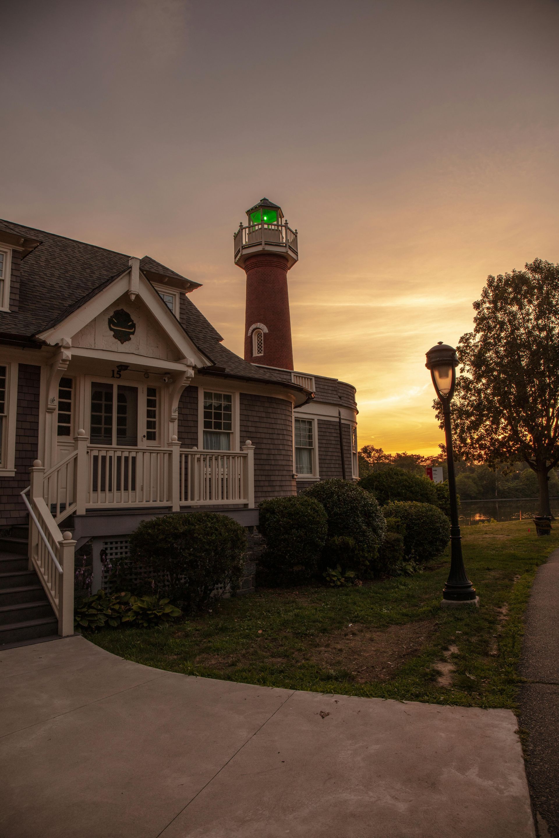 A brick lighthouse and building at sunset with an orange sky, green light, and street lamp.