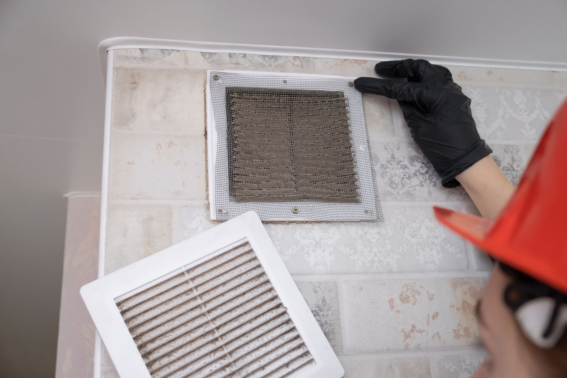 A person wearing gloves and a hard hat removes a dirty vent cover.