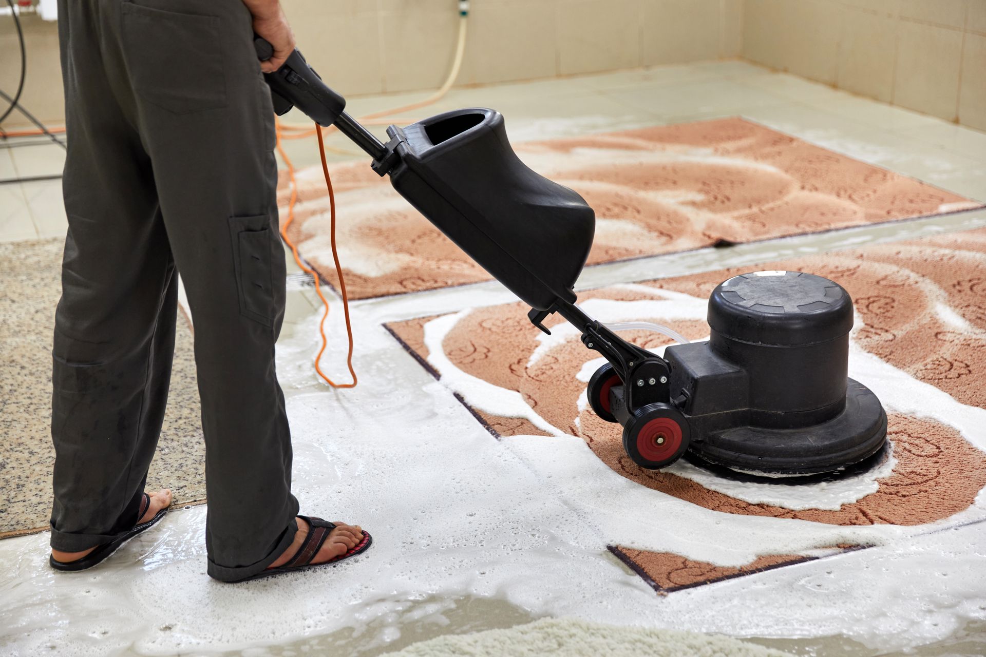 Person using a floor buffer to clean a rug in a room with a concrete floor.