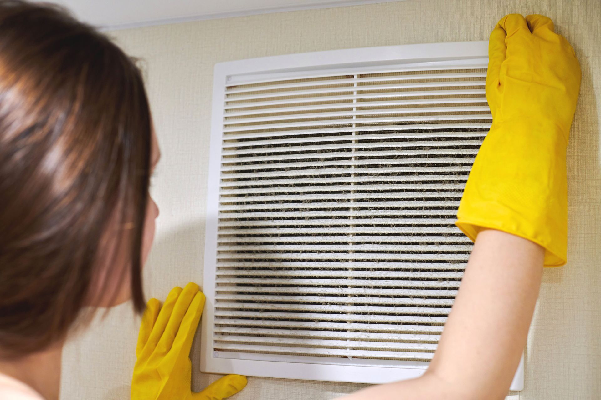 Person wearing yellow gloves cleans a dirty air vent on a wall.
