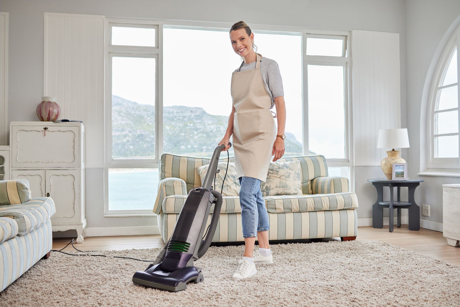 Woman vacuuming a rug in a living room, wearing an apron, smiling.