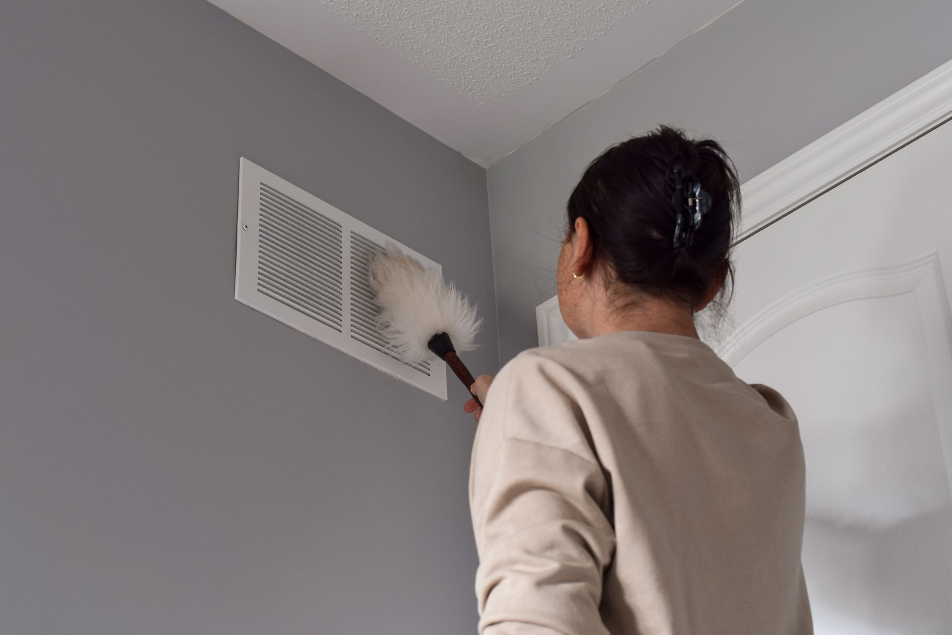 Person dusts a vent with a feather duster in a gray-walled room.