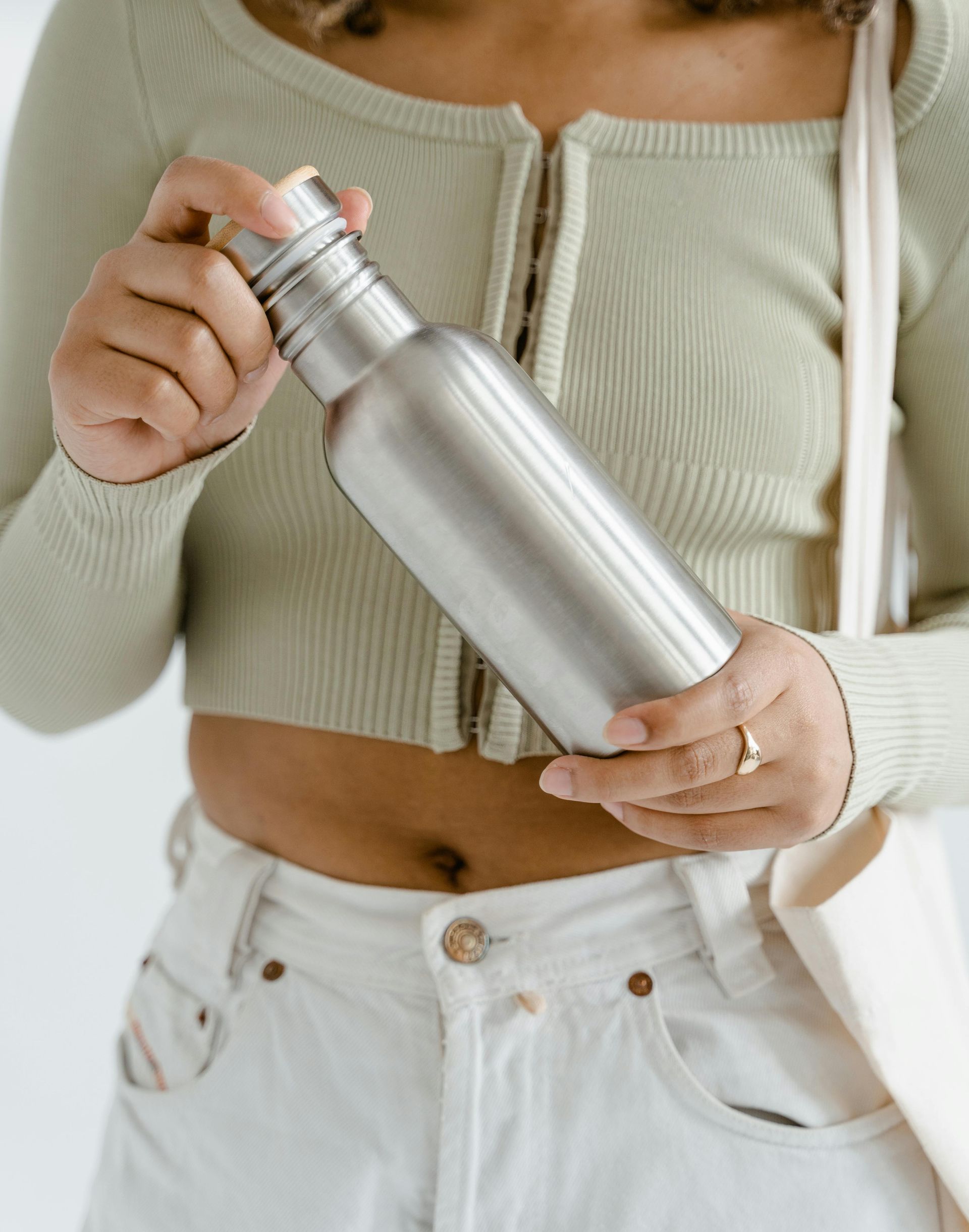 Vrouw met een zilverkleurige waterfles en een draagtas, gekleed in een groene top en een witte spijkerbroek.