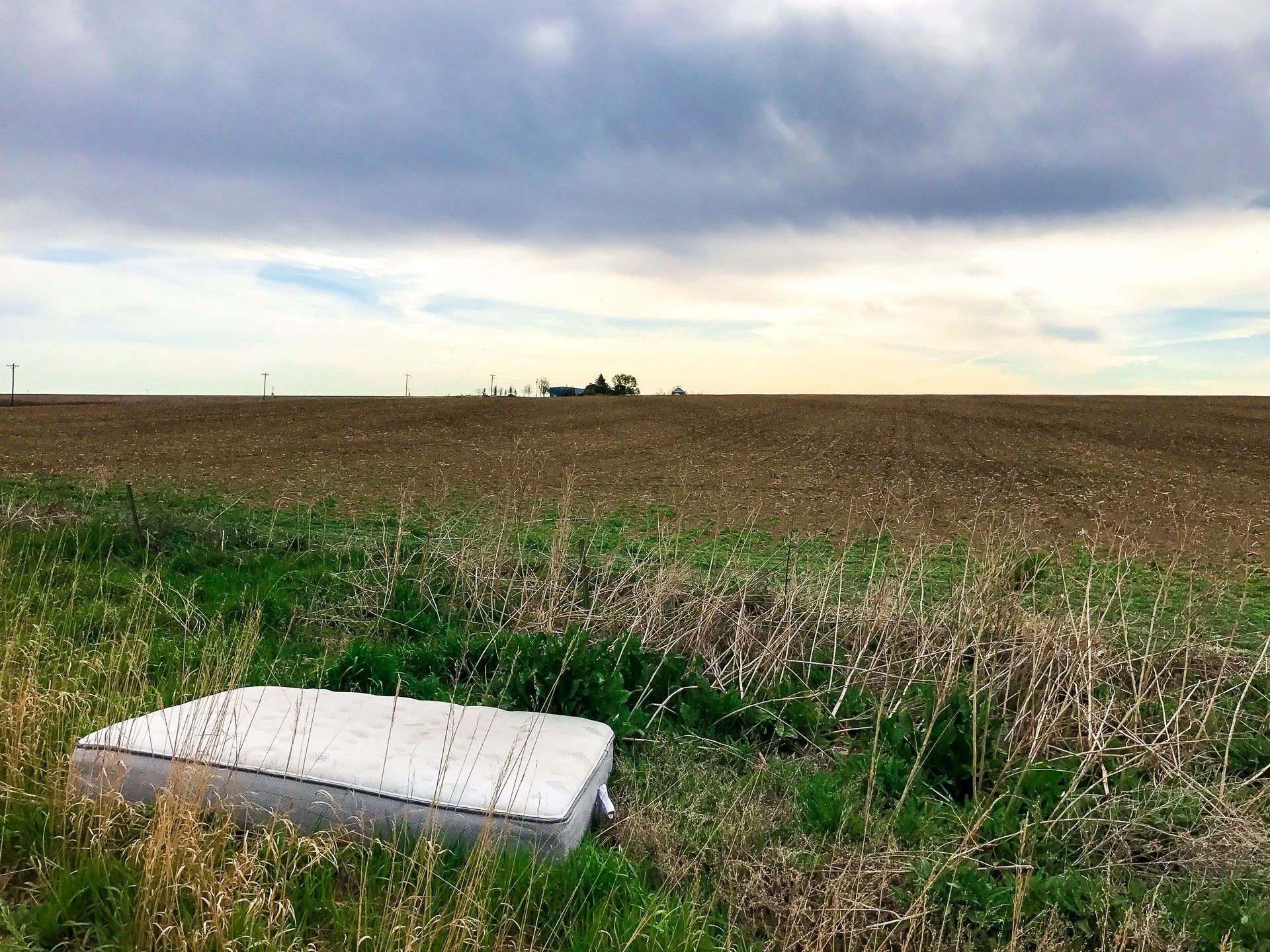 A white mattress lies in a grassy field under a cloudy sky.