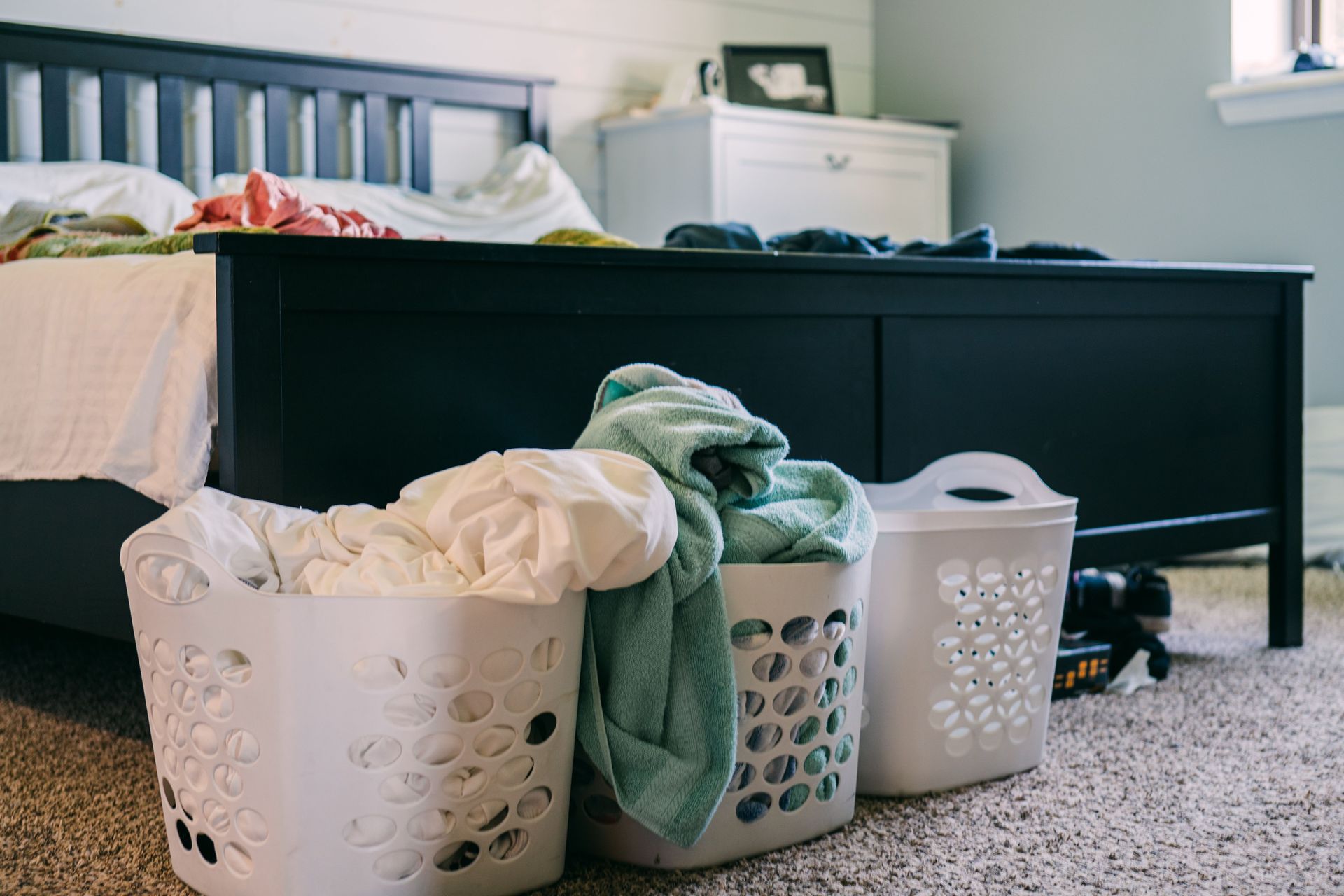 Laundry baskets of clothes in a bedroom beside a black bed frame.