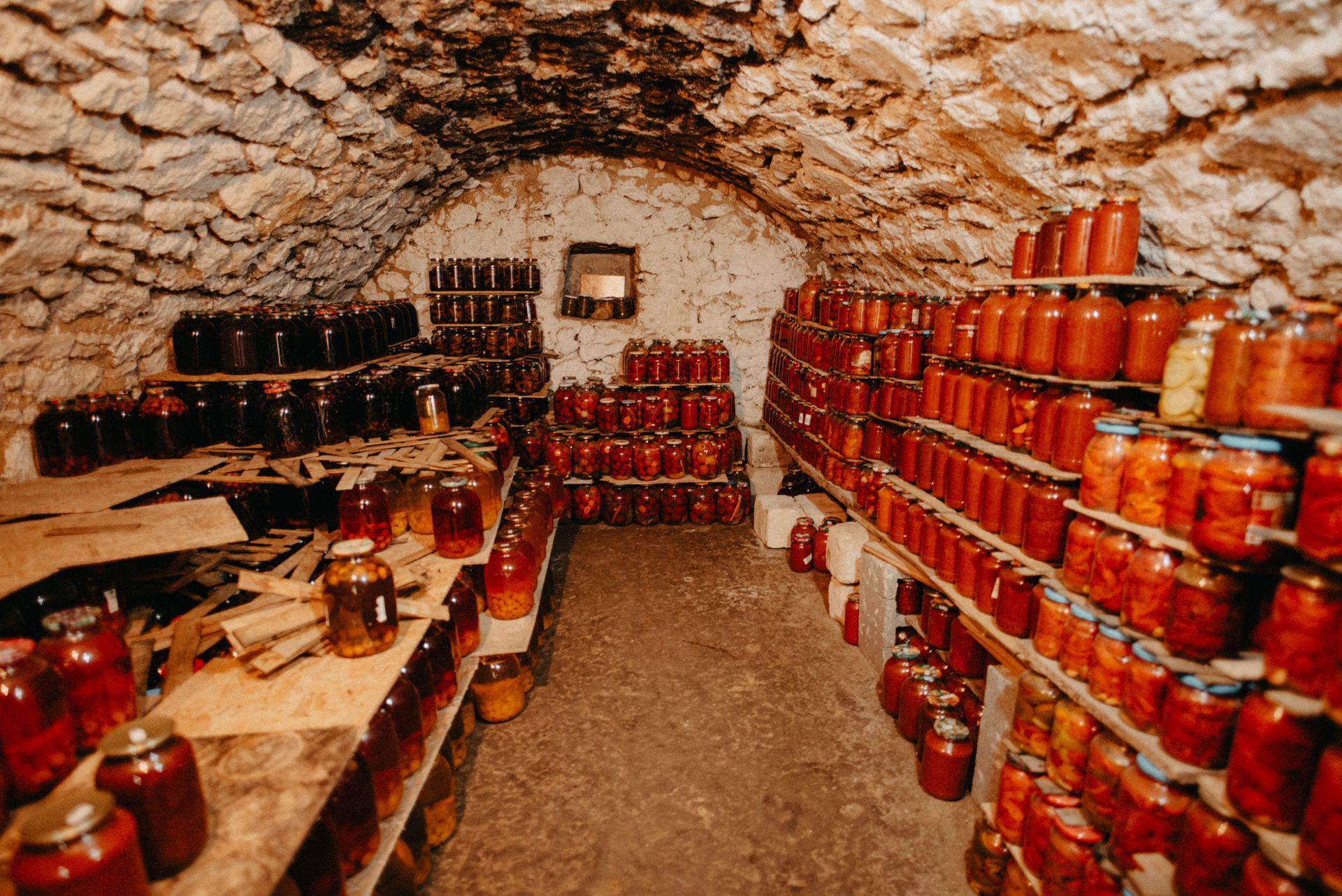 Stone cellar lined with shelves of red jars and bottles, with a rustic arched ceiling