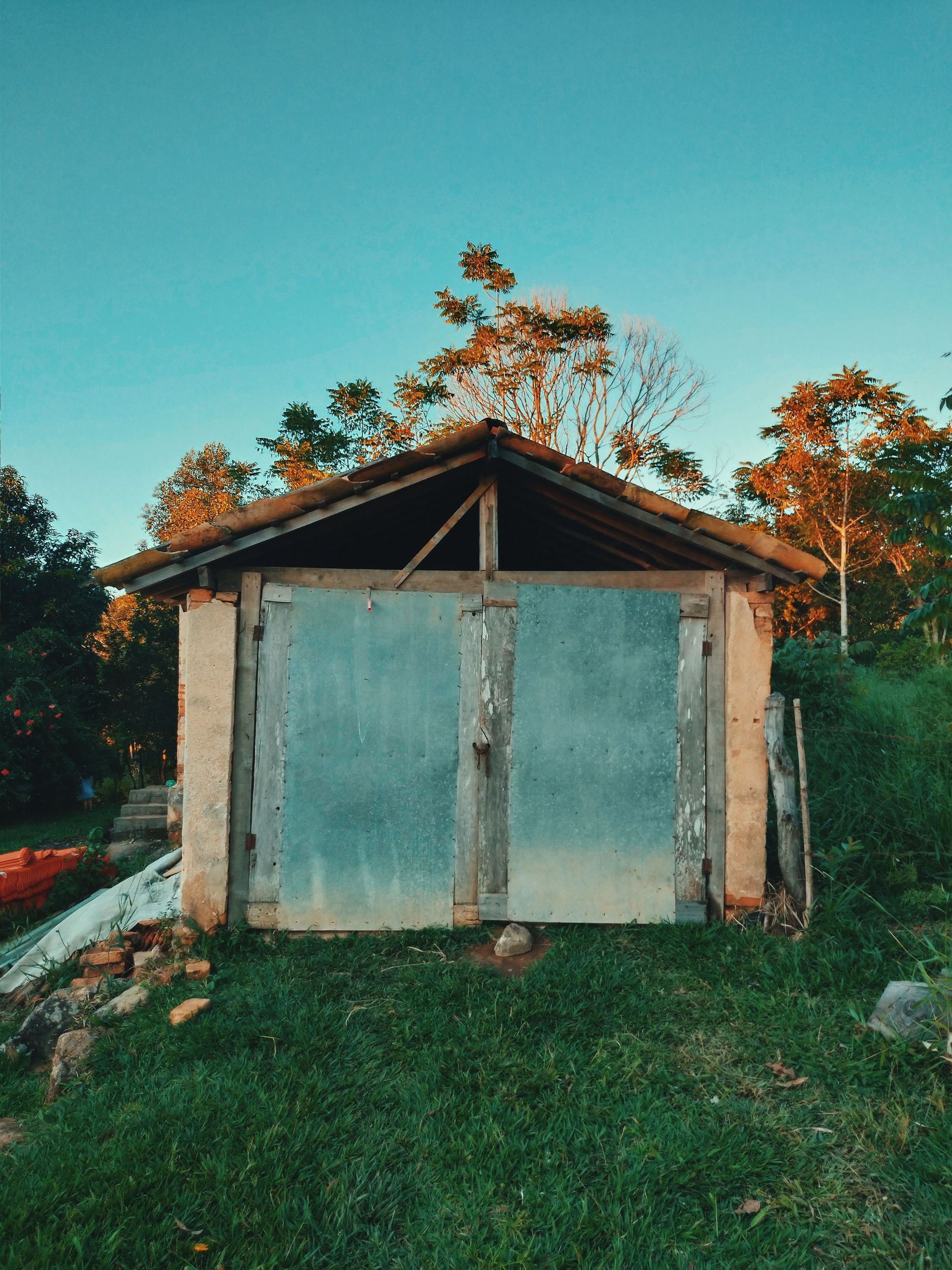 Small rustic shed with weathered metal doors, surrounded by grass and trees at sunset.