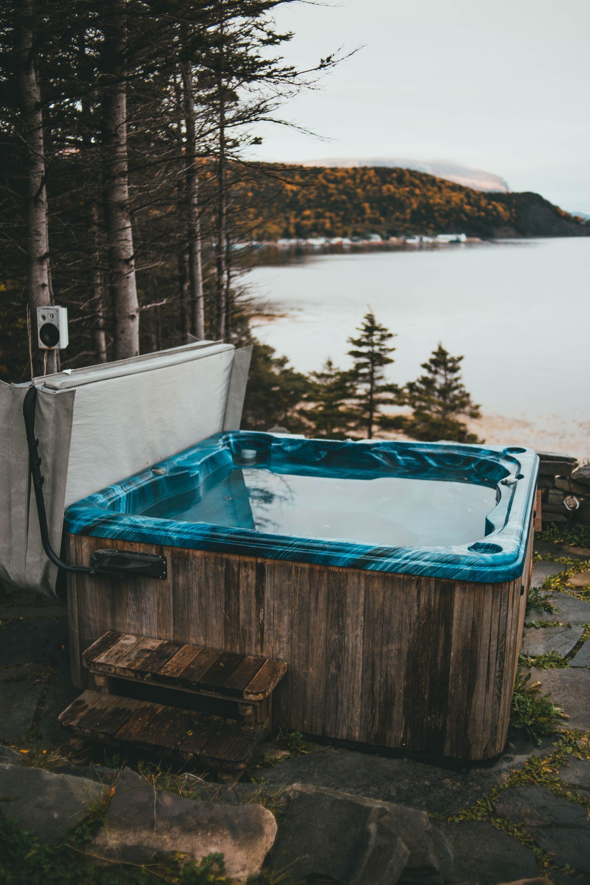 Wooden hot tub with blue water beside a lake, surrounded by trees at dusk